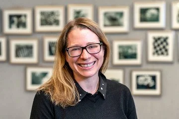 Young woman with glasses and long hair smiling, standing in front of a gallery wall with black and white framed photographs.