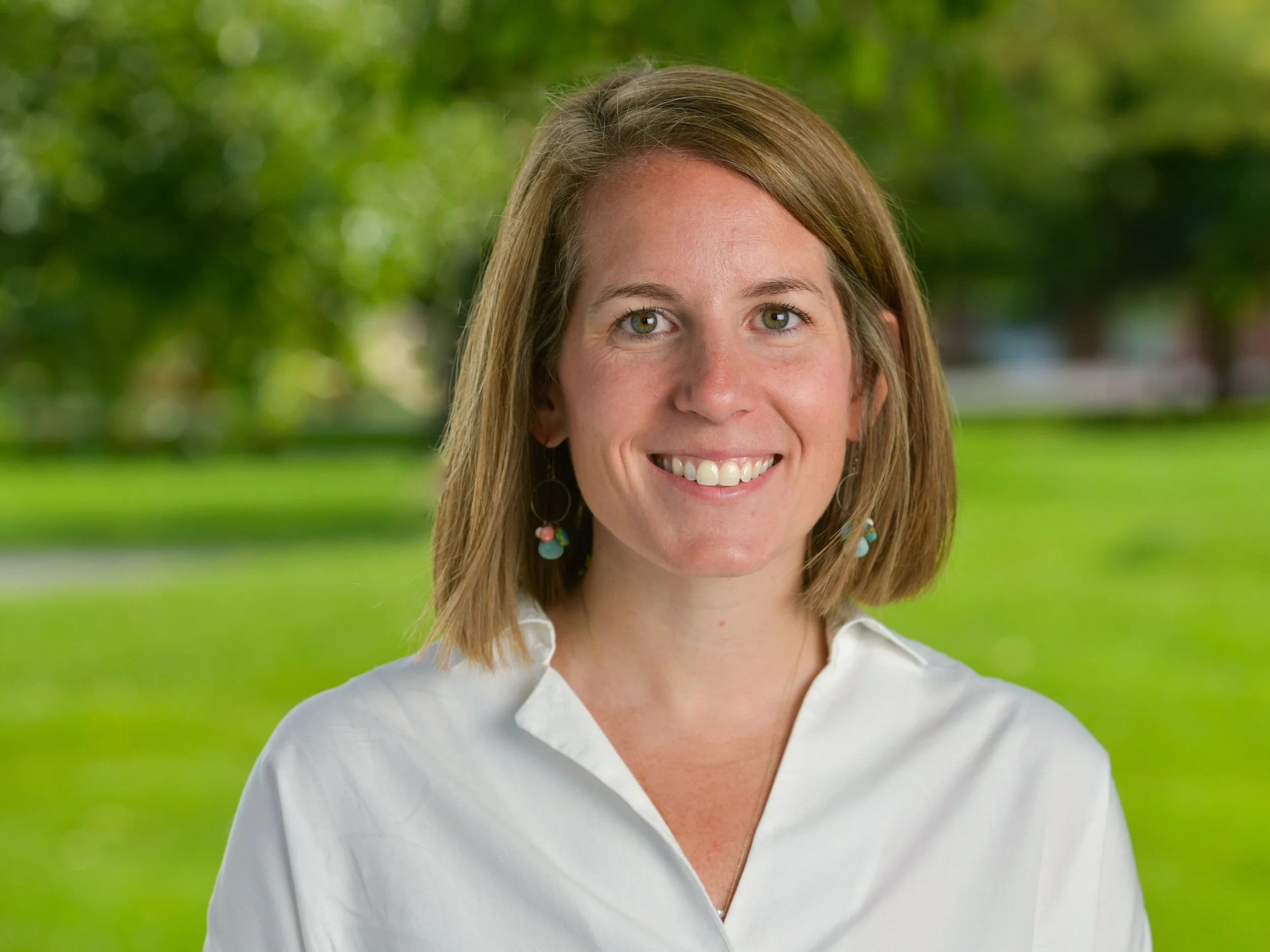 A smiling woman with shoulder-length brown hair, wearing a white shirt, standing outdoors with a blurred green park background.