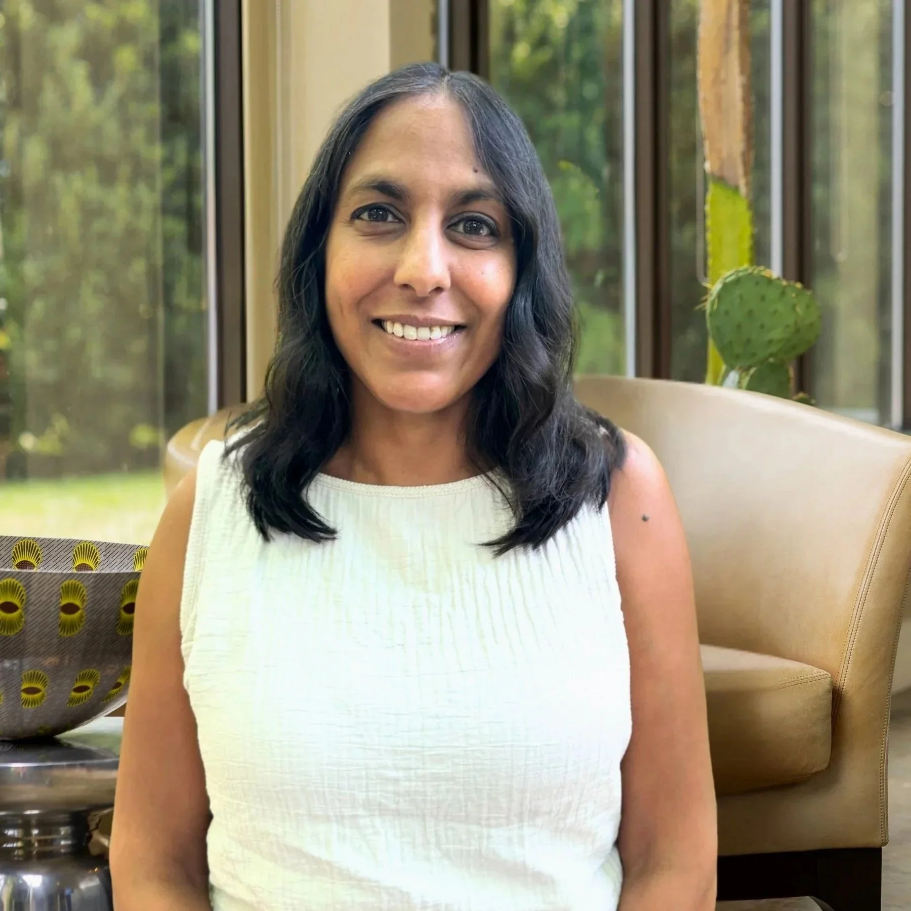 A woman with shoulder-length black hair wearing a sleeveless white top smiling indoors, with windows showing greenery outside and a cactus in the background.