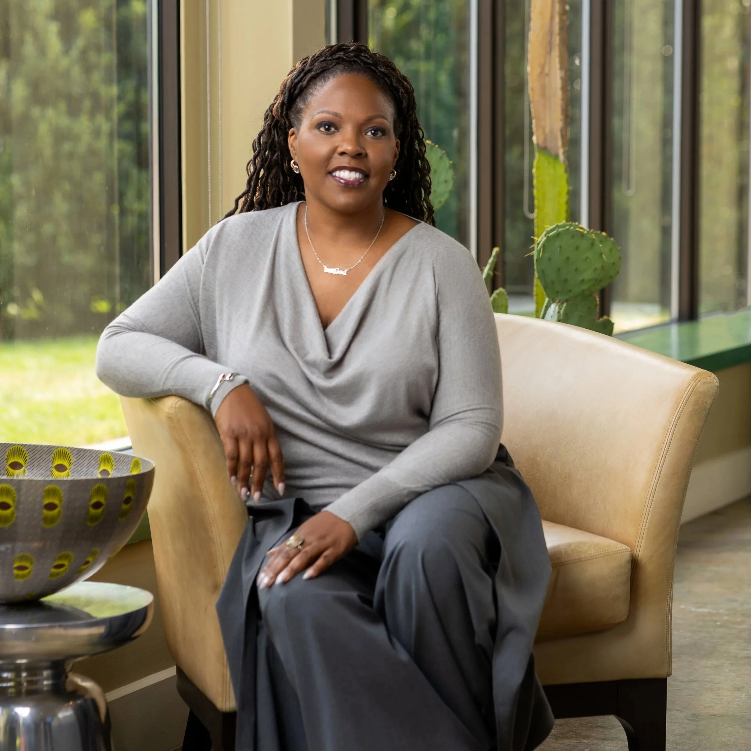 A woman sitting comfortably on a beige chair near a large window with cacti and greenery outside, wearing a gray top and dark pants.