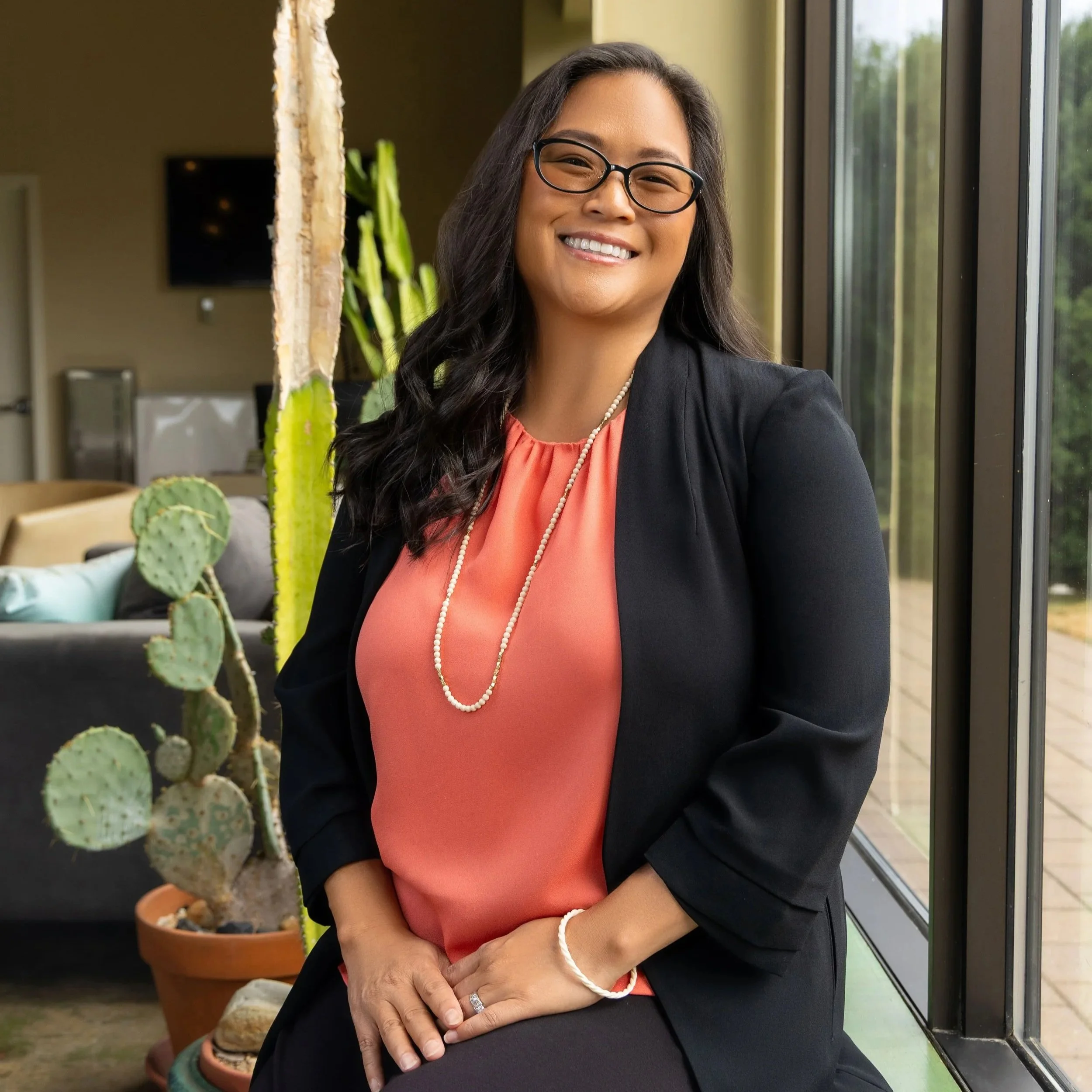 A woman with long black hair, glasses, and wearing a coral blouse with a black blazer, smiling and sitting by a large window inside a modern office or lobby area, with cacti plants visible in the background.