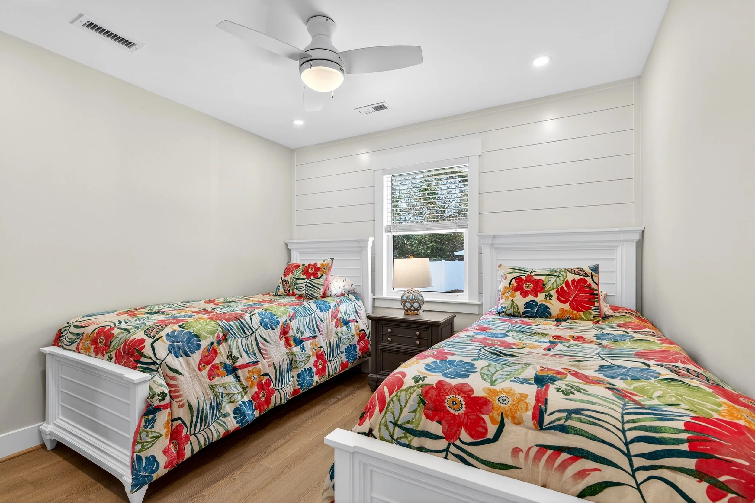 A bedroom with two twin beds featuring matching floral bedding, a central nightstand with a lamp, a window with white blinds, a ceiling fan, and wood flooring. Oak Island NC real estate photography. 