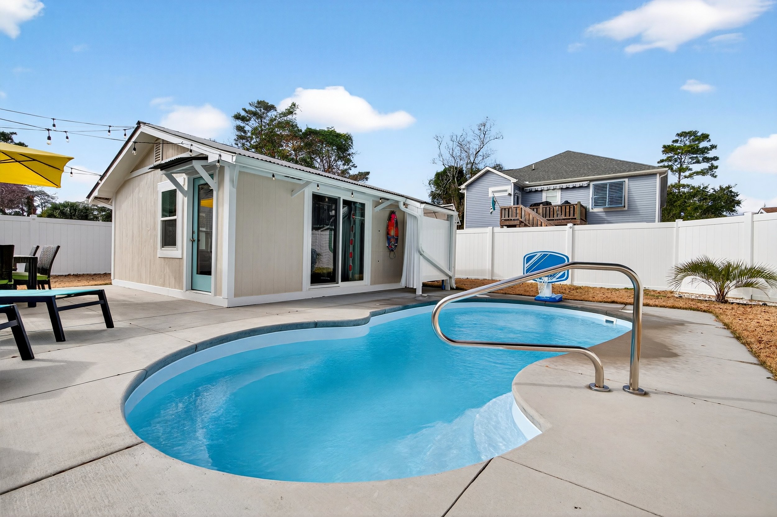 Backyard with small swimming pool, pool house, patio furniture, and a children's basketball hoop, surrounded by a white fence under a partly cloudy sky. Backyard pool at Oak Island beach house photographed for property listing