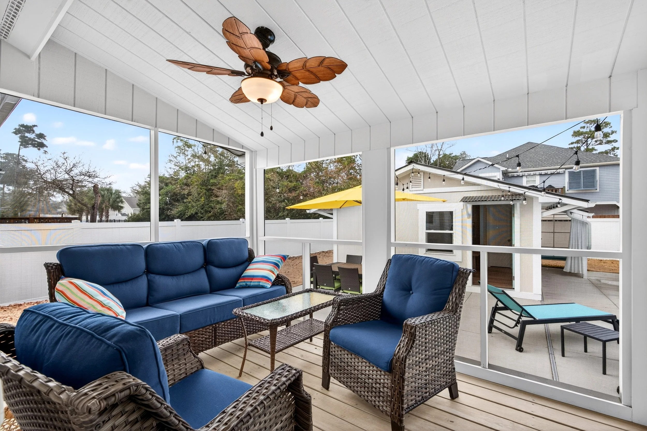 Enclosed porch with blue outdoor furniture, wicker chairs with blue cushions, a glass coffee table, ceiling fan with leaf-shaped blades, string lights, and a view of neighboring houses with patio umbrellas.