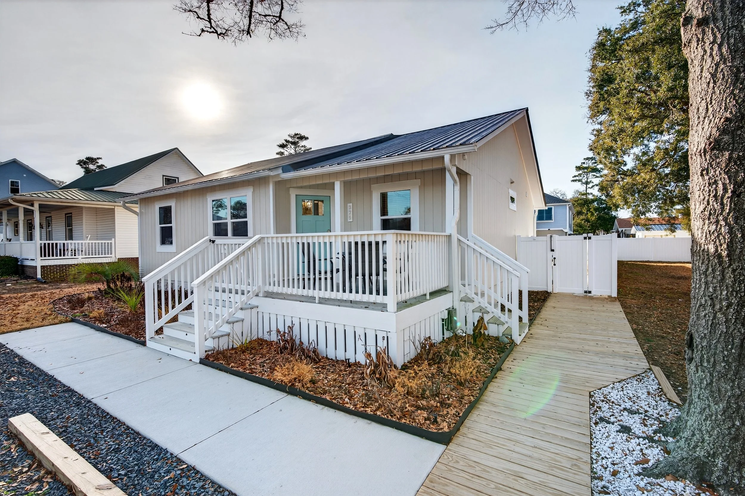 Front view of a white house with a front porch, green door, and a metal roof, surrounded by pathways, trees, and neighboring houses in a neighborhood. Oak Island beach house rental real estate listing photography.
