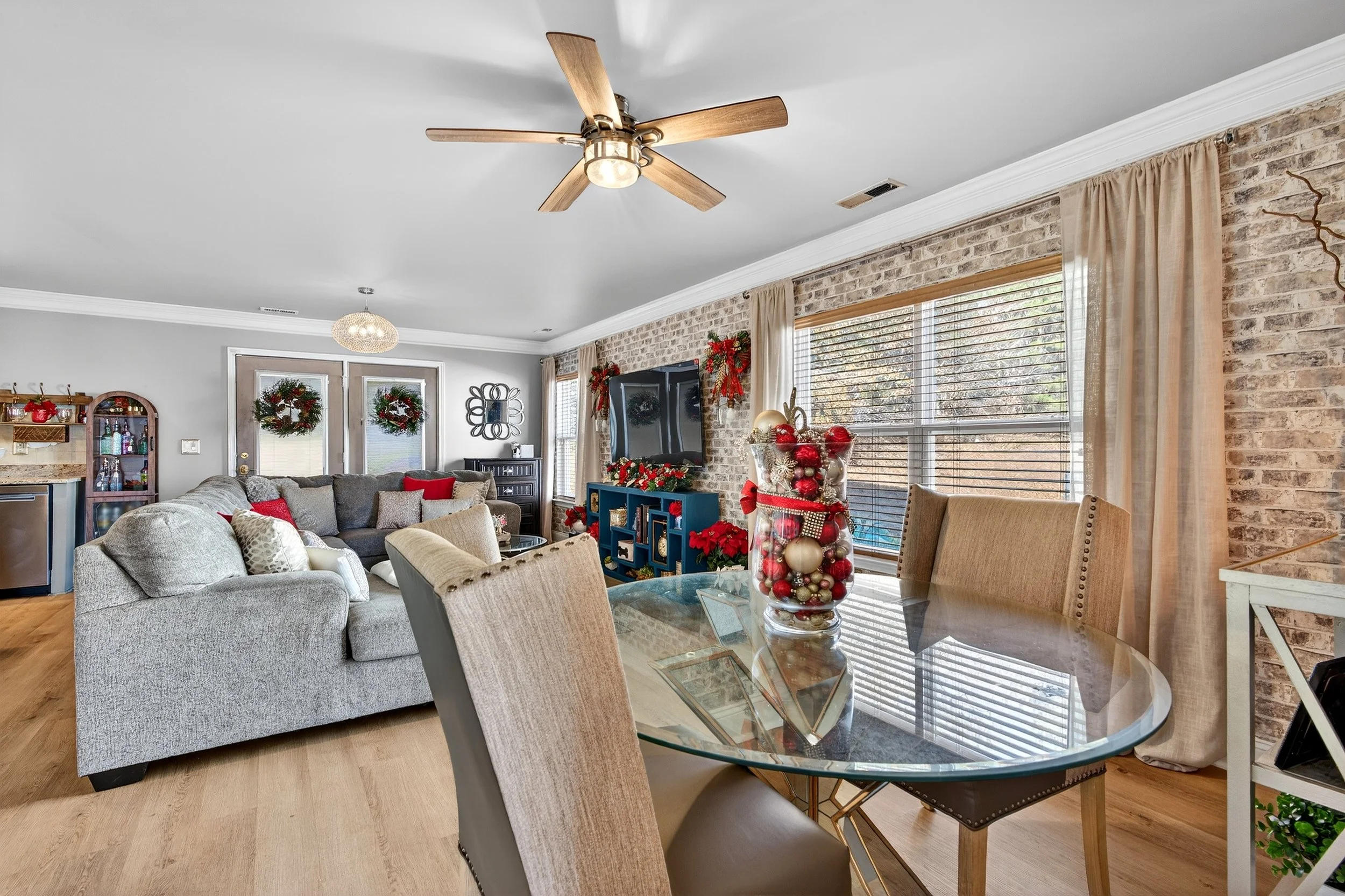 Living room decorated for Christmas with a gray sectional sofa, holiday ornaments, wreaths, and a flat-screen TV on a blue stand near a brick wall, with a glass dining table and beige chairs, and large windows with beige curtains.