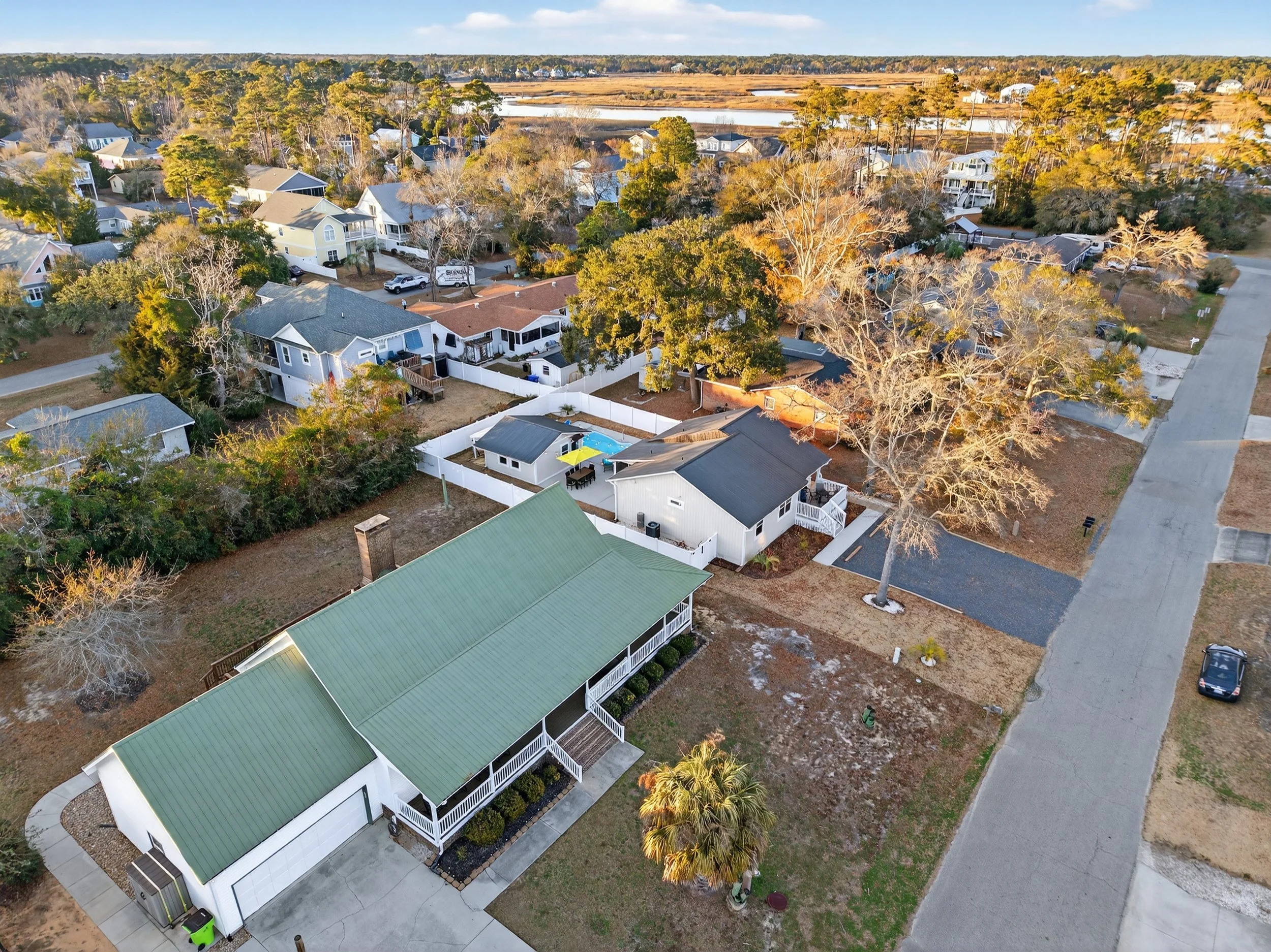 Aerial view of a residential neighborhood with various houses, trees, and yards; some homes have pools or decks, and a large green-roofed building is prominent in the foreground. Pool and outdoor living area at Oak Island beach home drone photo