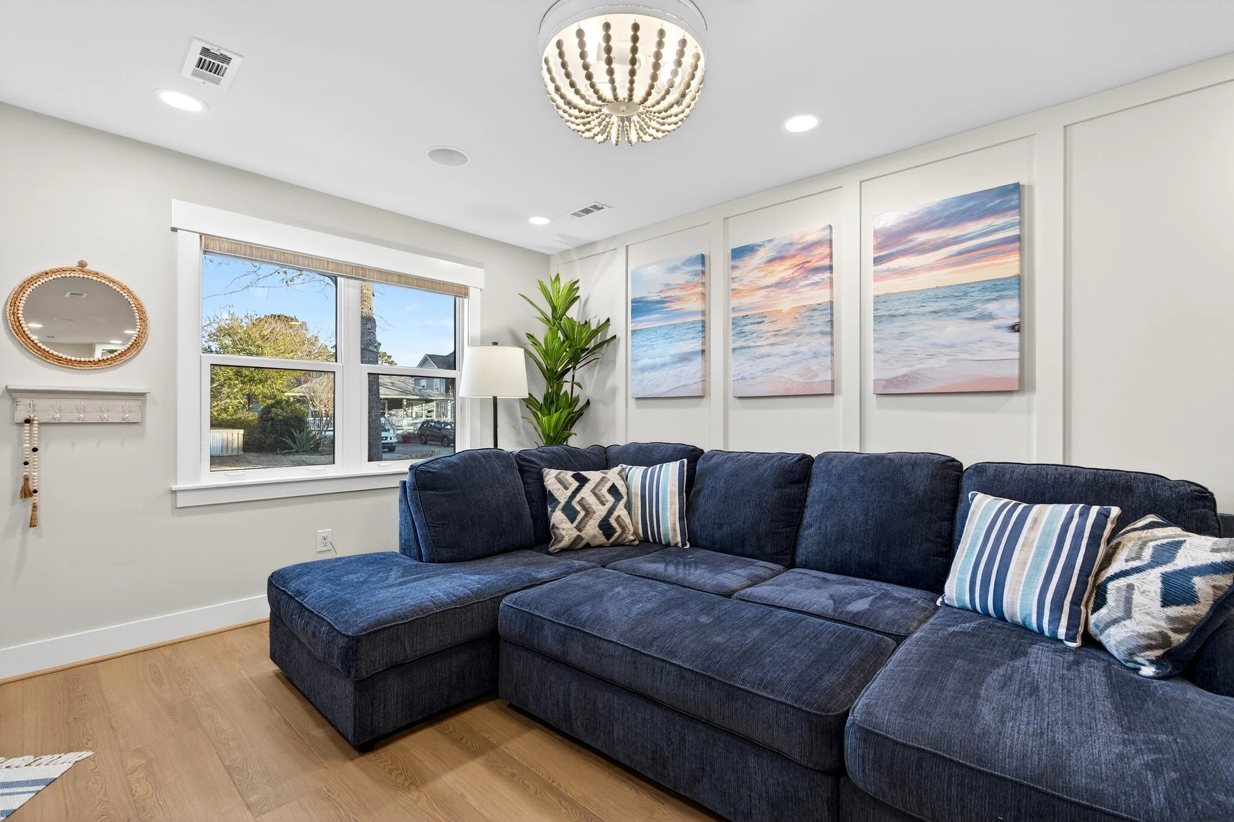 Living room with a dark blue sectional sofa, decorative pillows, a large window, framed marine-themed artwork, a potted plant, a standing lamp, and a ceiling light fixture. Open living room in Oak Island beach home photographed for real estate market