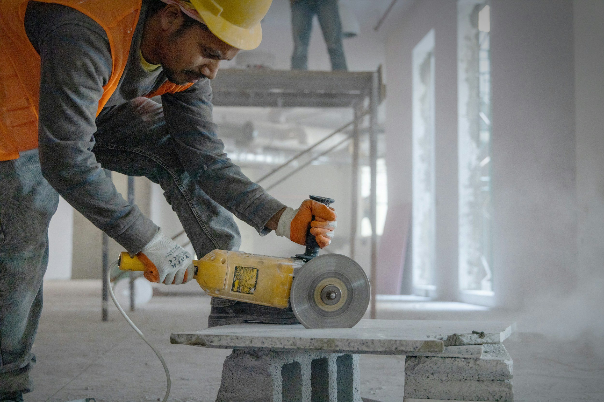 A construction worker wearing a yellow hard hat, orange safety vest, and gray clothes is using a handheld power saw to cut a piece of material on a table in a building under construction.