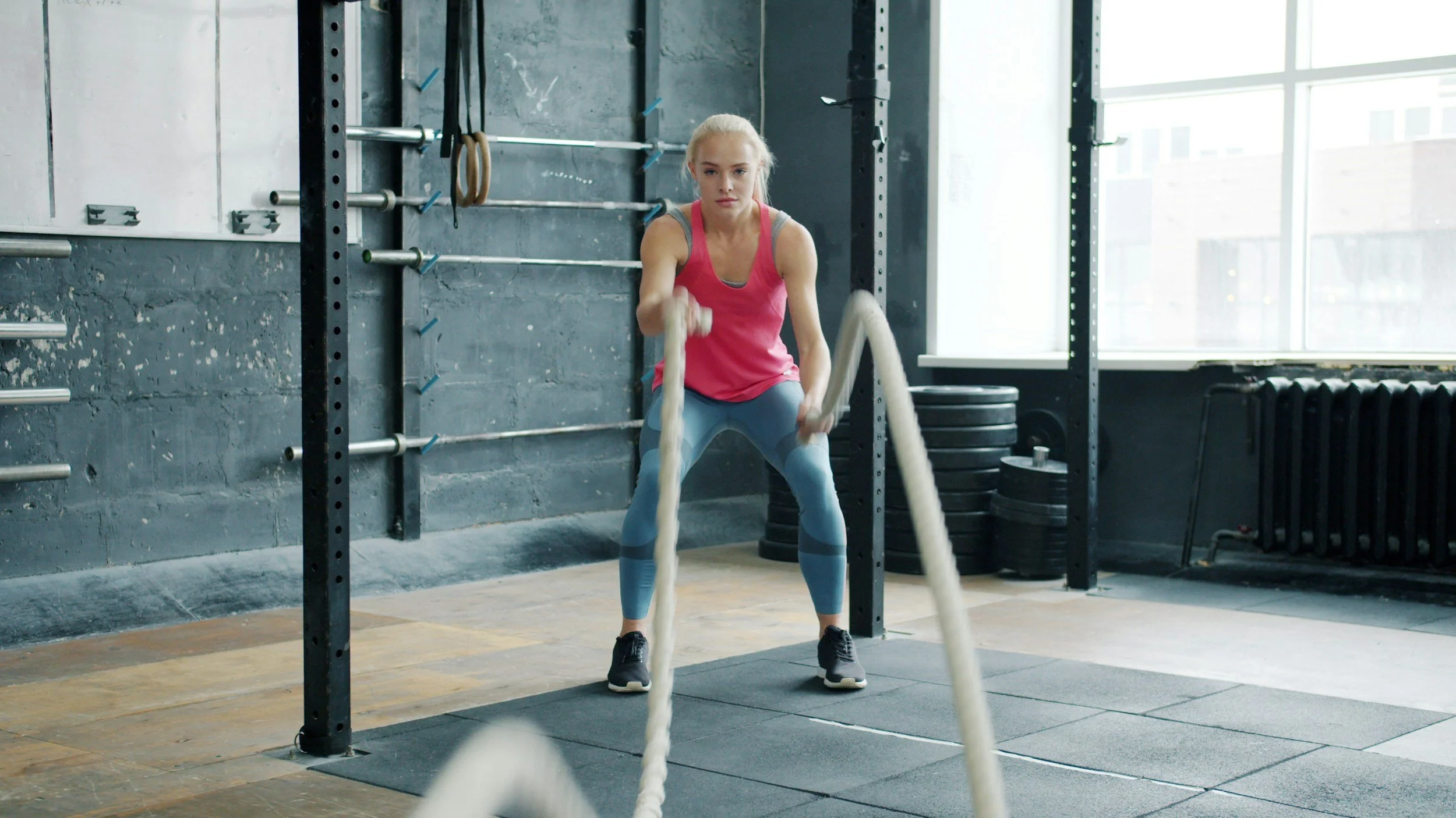 A woman in a pink tank top and blue workout leggings exercises with battle ropes inside a gym.