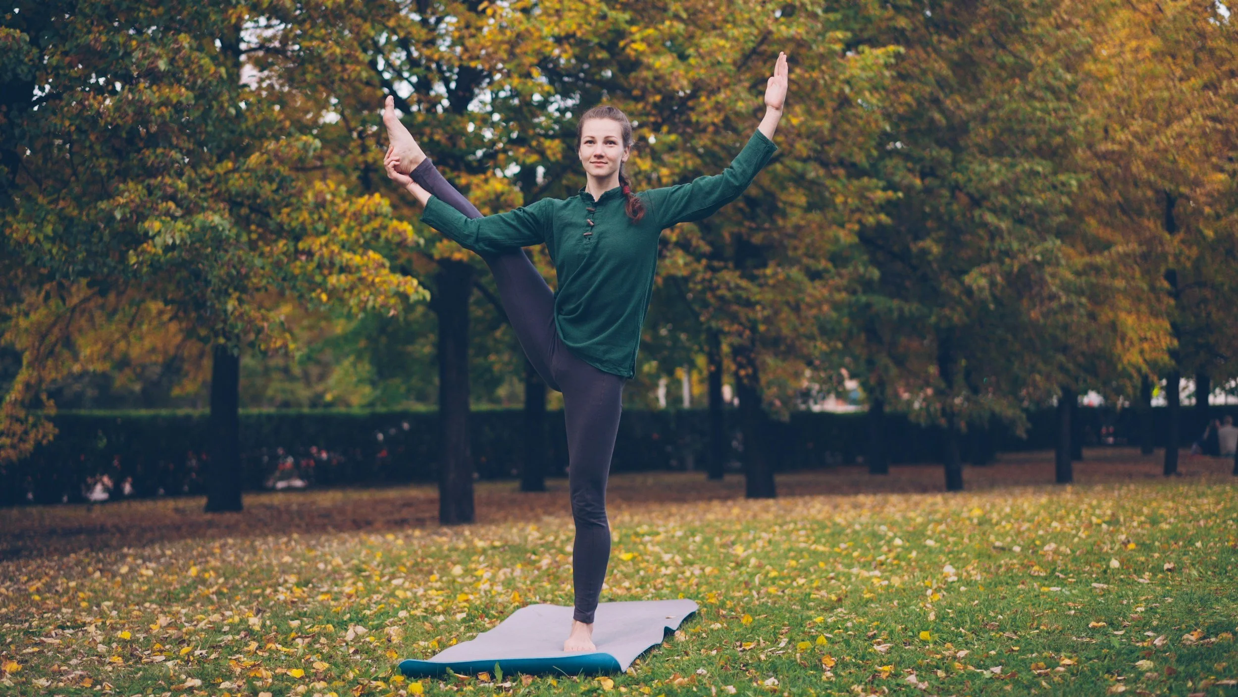 A woman practicing yoga outdoors on a fall day, balancing on one leg with one arm raised and the other holding her raised foot behind her, standing on a yoga mat in a park with autumn leaves and trees in the background.
