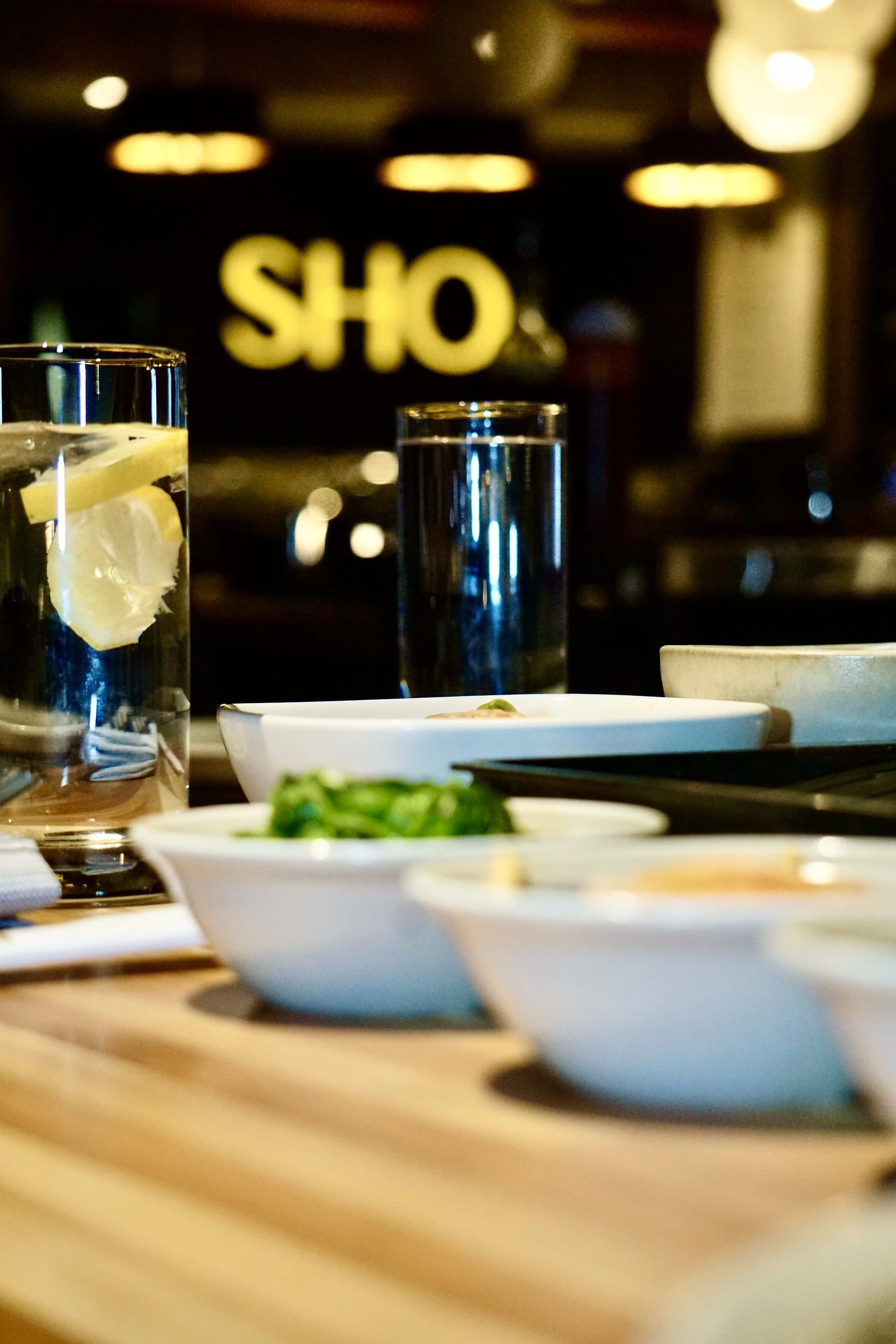 Close-up of a dining table with bowls of food and glasses of water, with a blurred background of a restaurant or bar displaying a sign that reads 'SHO'.