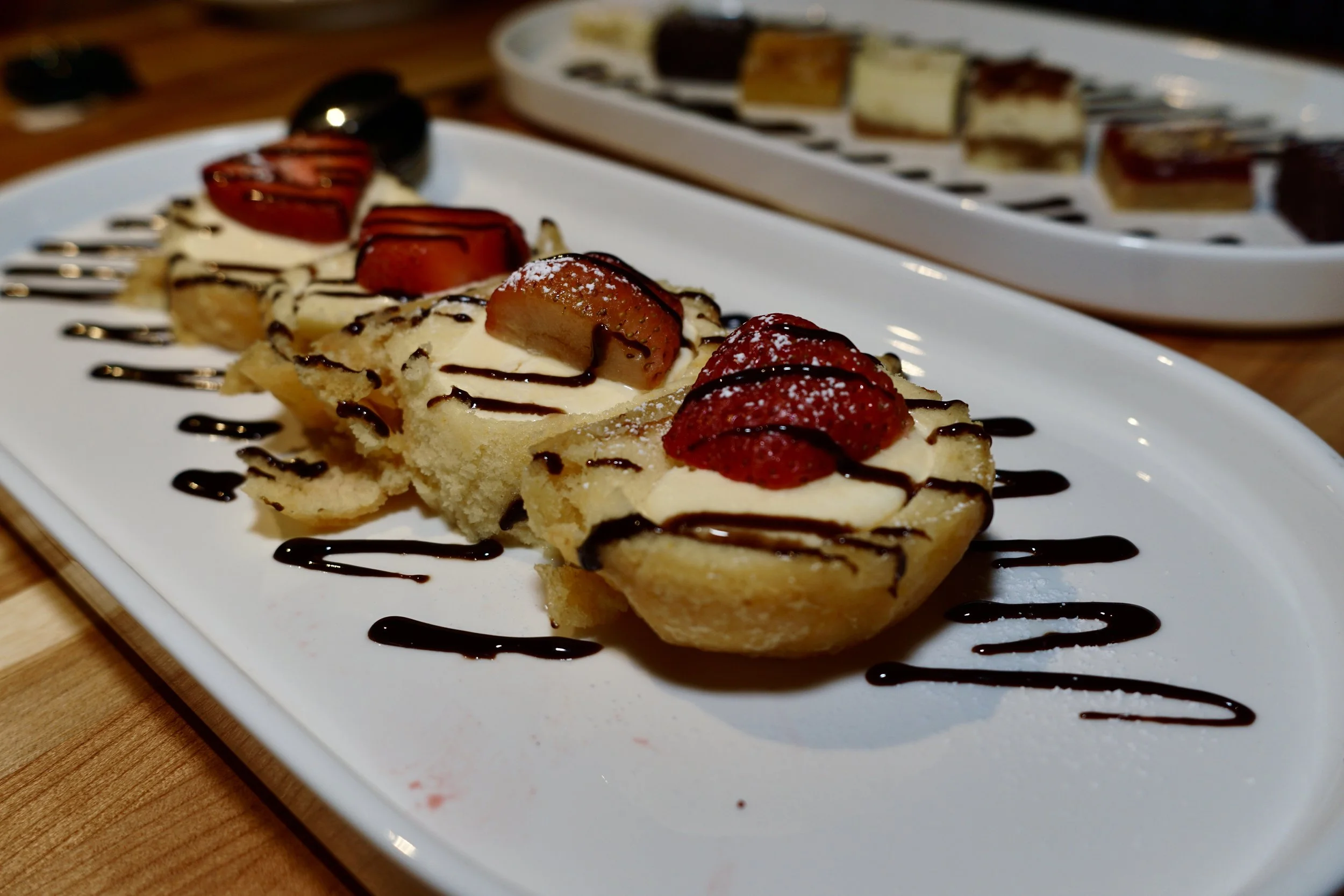 Three small dessert tartlets with sliced strawberries, whipped cream, and chocolate drizzle, garnished with powdered sugar, on a white rectangular plate.