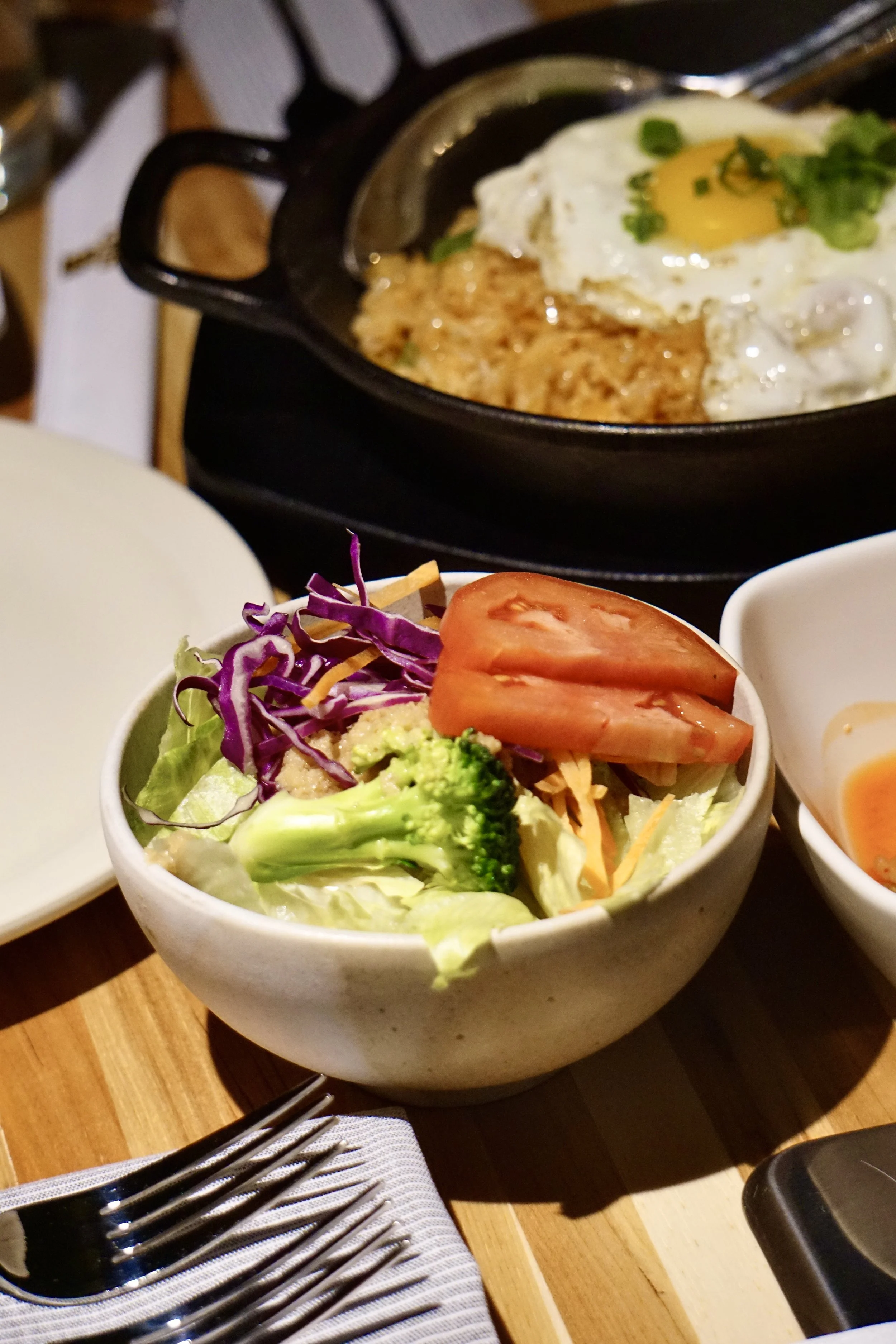 Side salad with lettuce, purple cabbage, shredded cheese, tomato slices, and broccoli in a white bowl, with a fork and napkin nearby, and a skillet with a fried egg, green onion, and hash browns in the background.