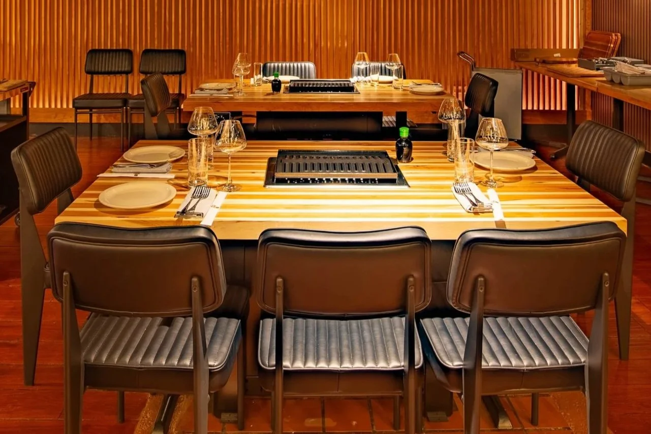 A modern dining room table set with plates, glasses, and cutlery, with a built-in grill in the center, surrounded by black cushioned chairs, and wooden walls in the background.
