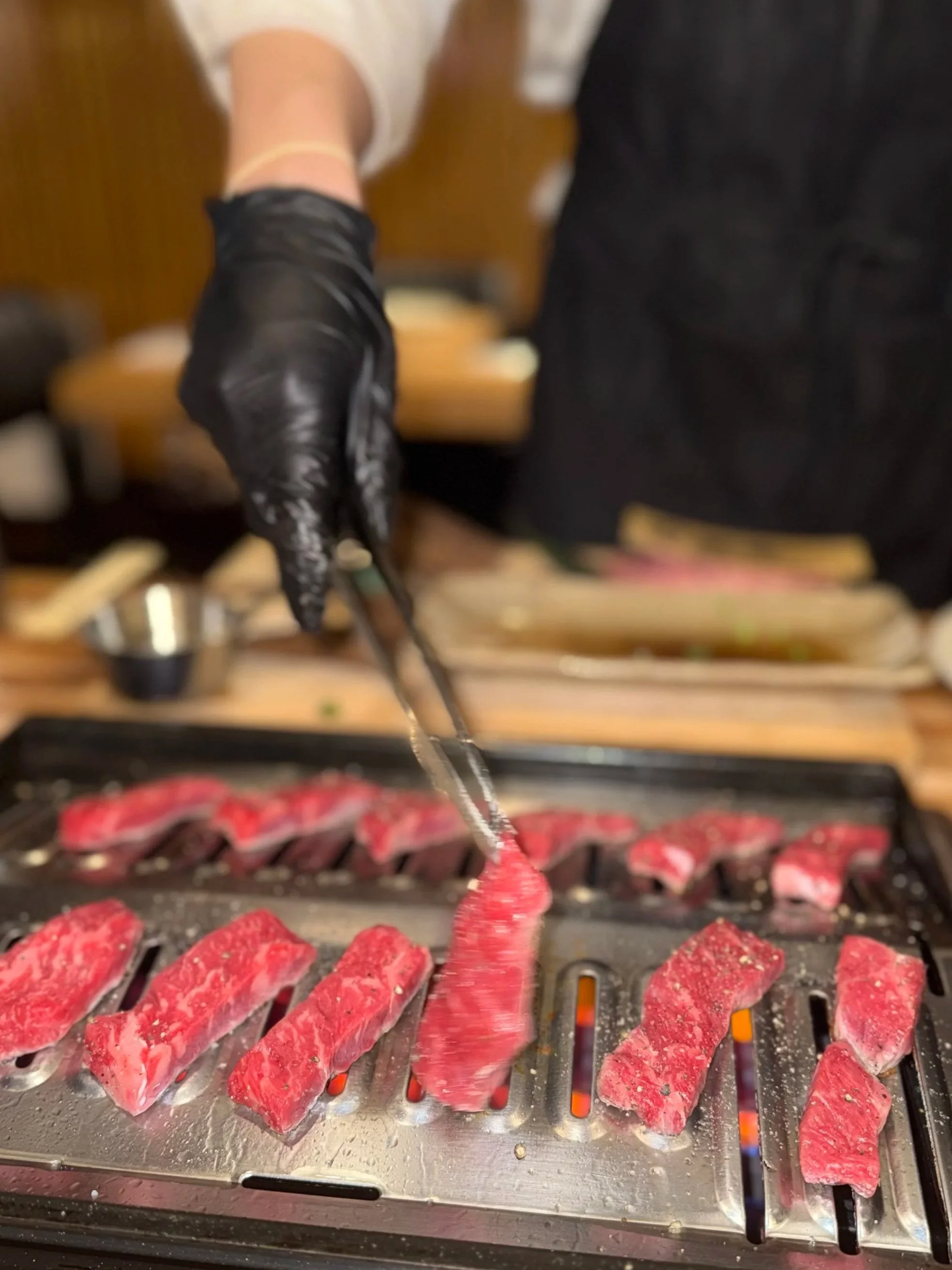 A person wearing black gloves using tongs to grill pieces of raw red meat on a flat grill, with a blurred background of a wooden table and kitchen setting.
