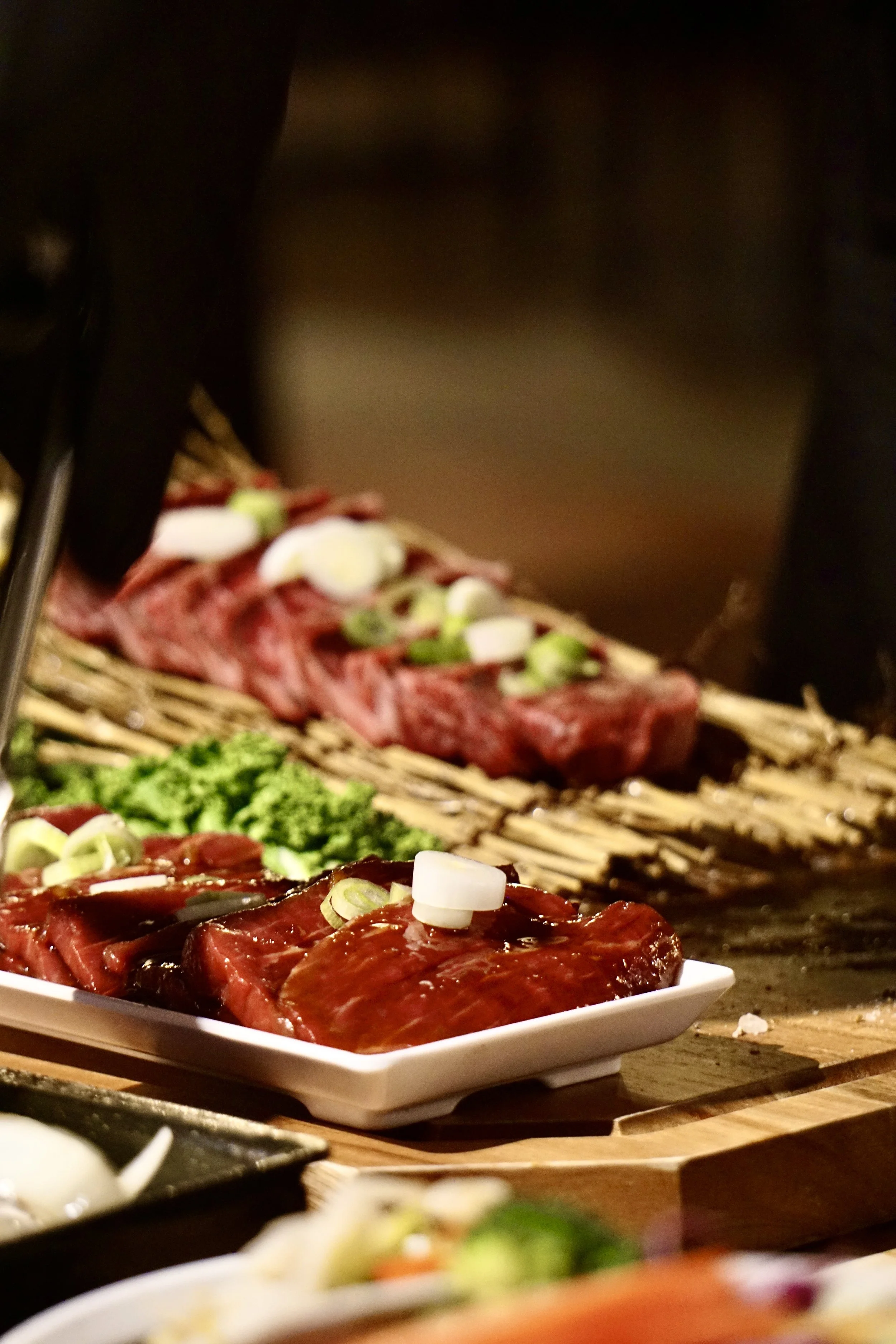 Close-up of raw marinated beef and vegetables on plates, with skewered meat in the background at a barbecue or grill.