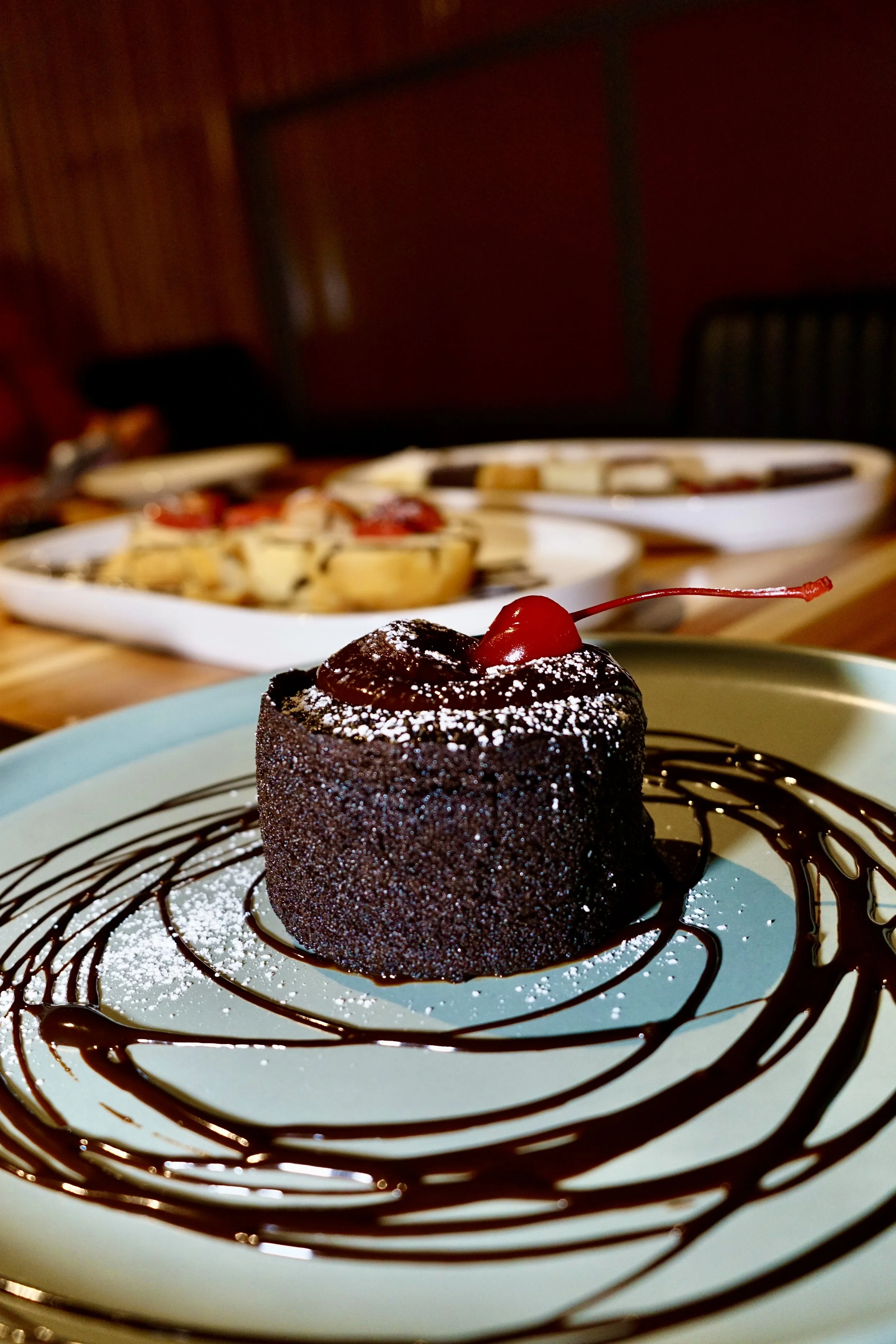 A chocolate lava cake garnished with a cherry on top, dusted with powdered sugar, and drizzled with chocolate syrup on a white plate.