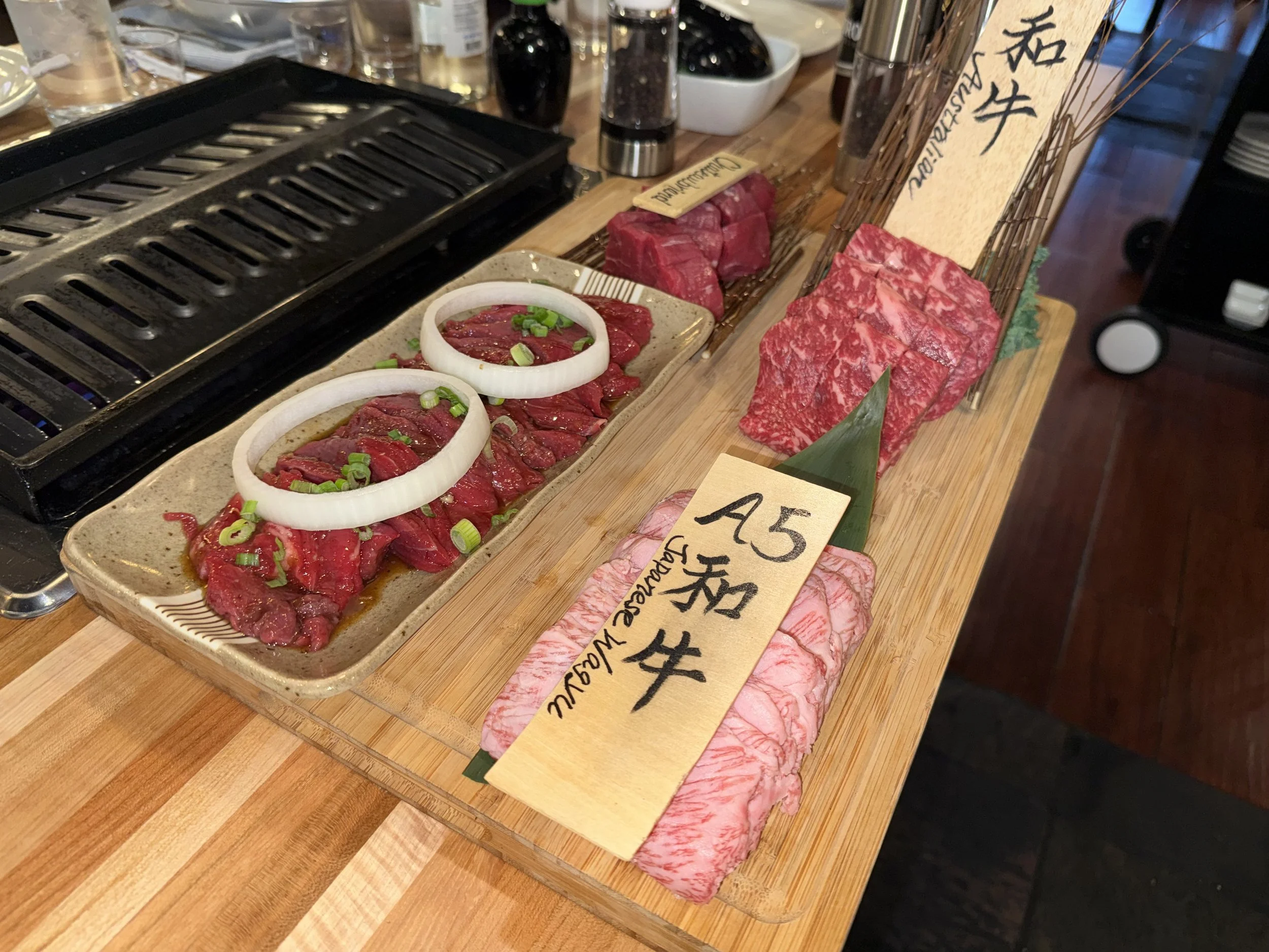 Display of various cuts of marbled beef with handwritten labels on a wooden board at a Japanese restaurant.