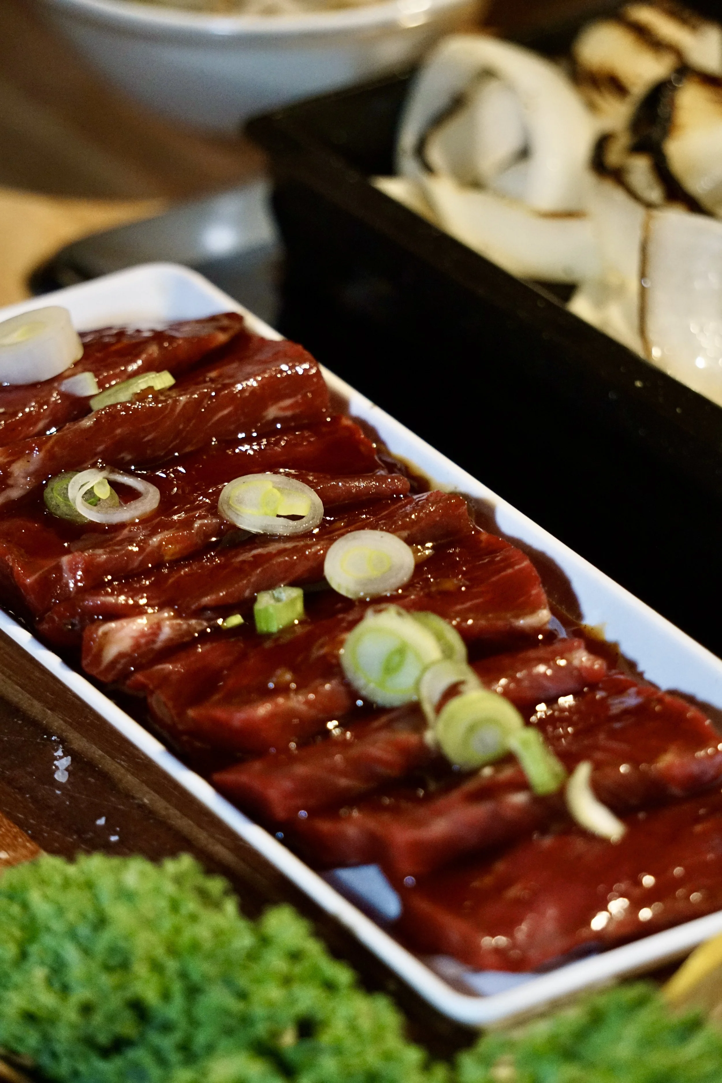 Thinly sliced raw beef marinated in sauce, garnished with sliced green onions, served on a white rectangular plate with grilled vegetables in the background.