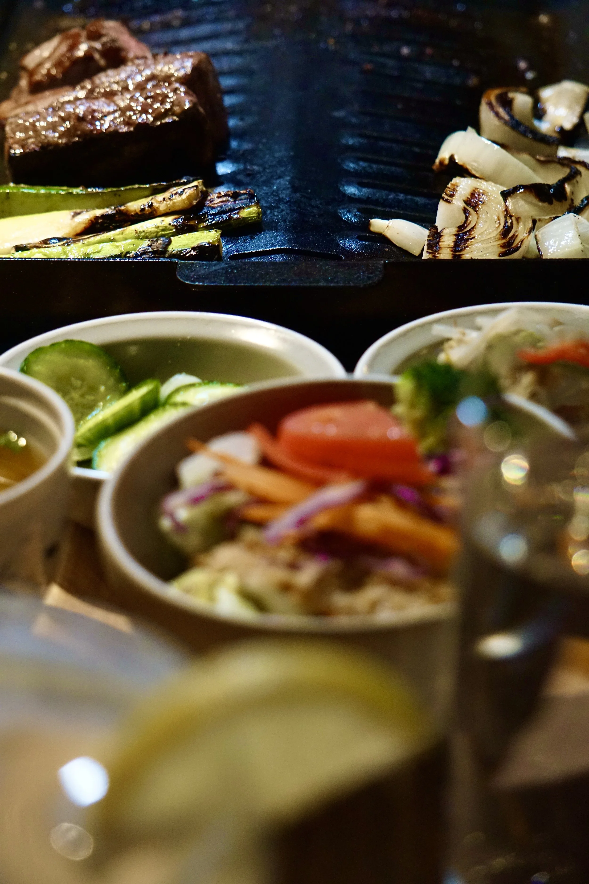 Grilled vegetables, steak, and small bowls of salad with cucumbers, tomatoes, and shredded vegetables on a barbecue grill.