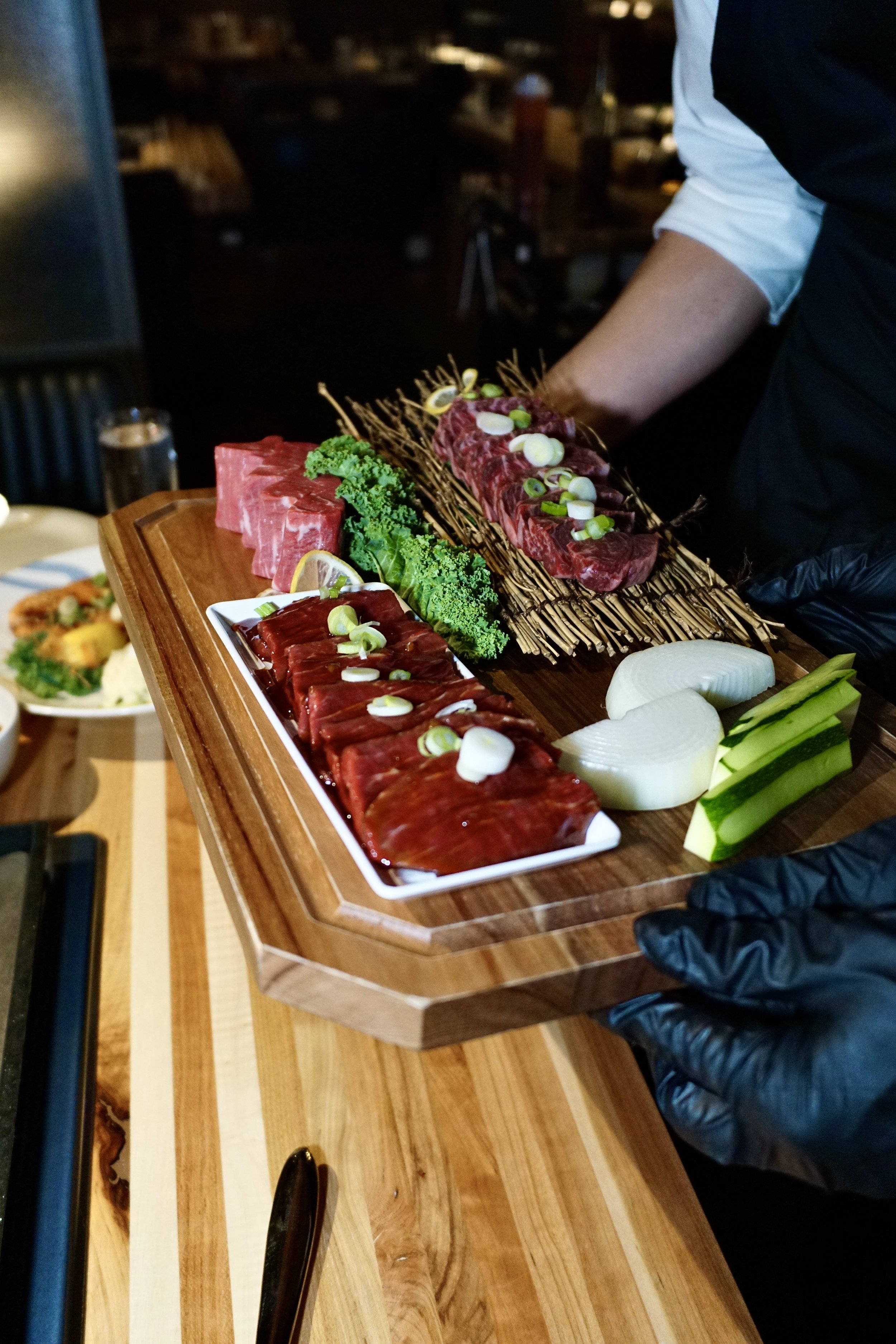 A person wearing black gloves holds a wooden platter with various raw cuts of beef, garnished with green onions, lemon slices, and fresh vegetables.