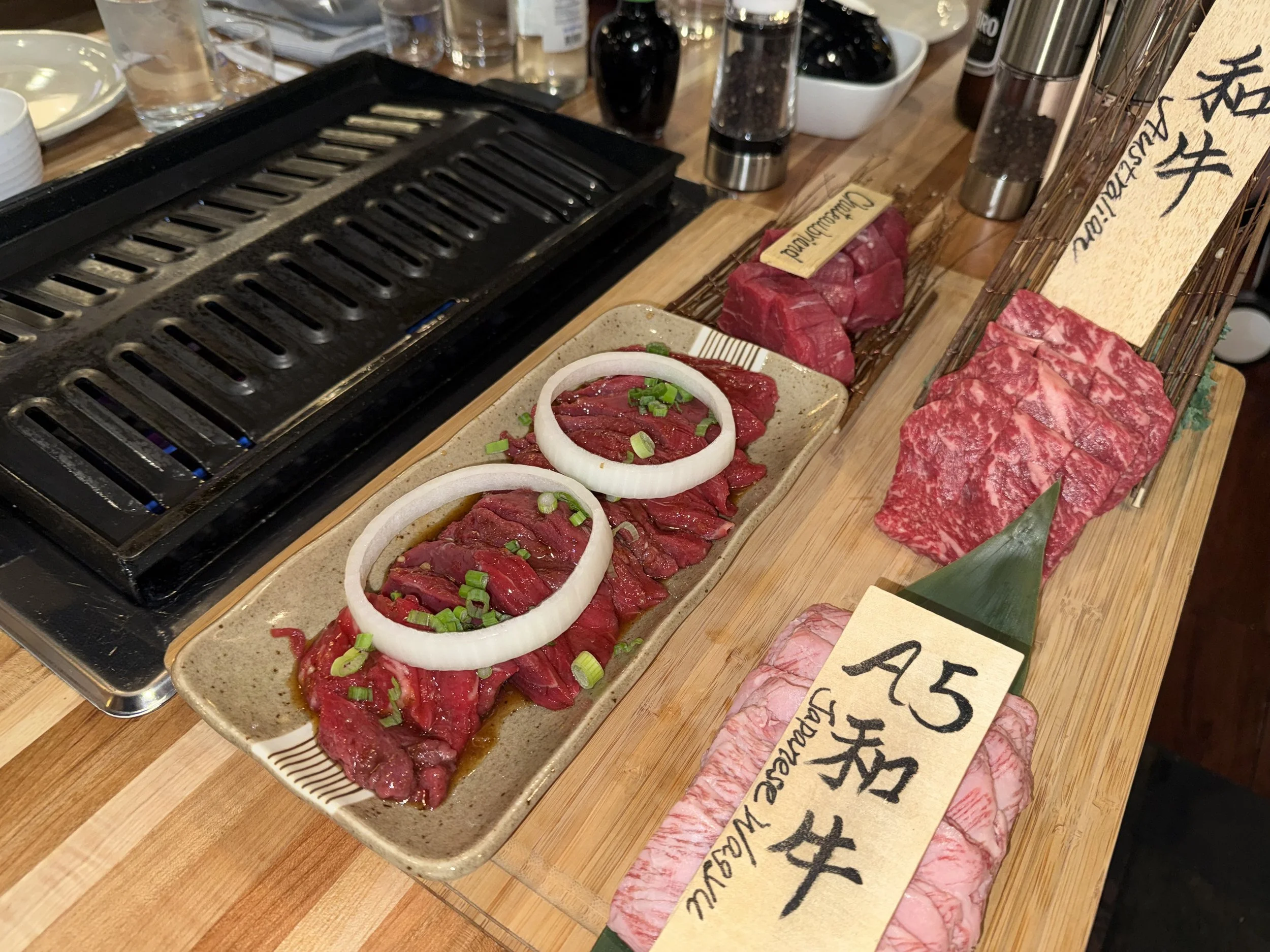 Various cuts of raw beef on a butcher's board, with some topped with sliced onions and green onions, ready for grilling at a Japanese restaurant.