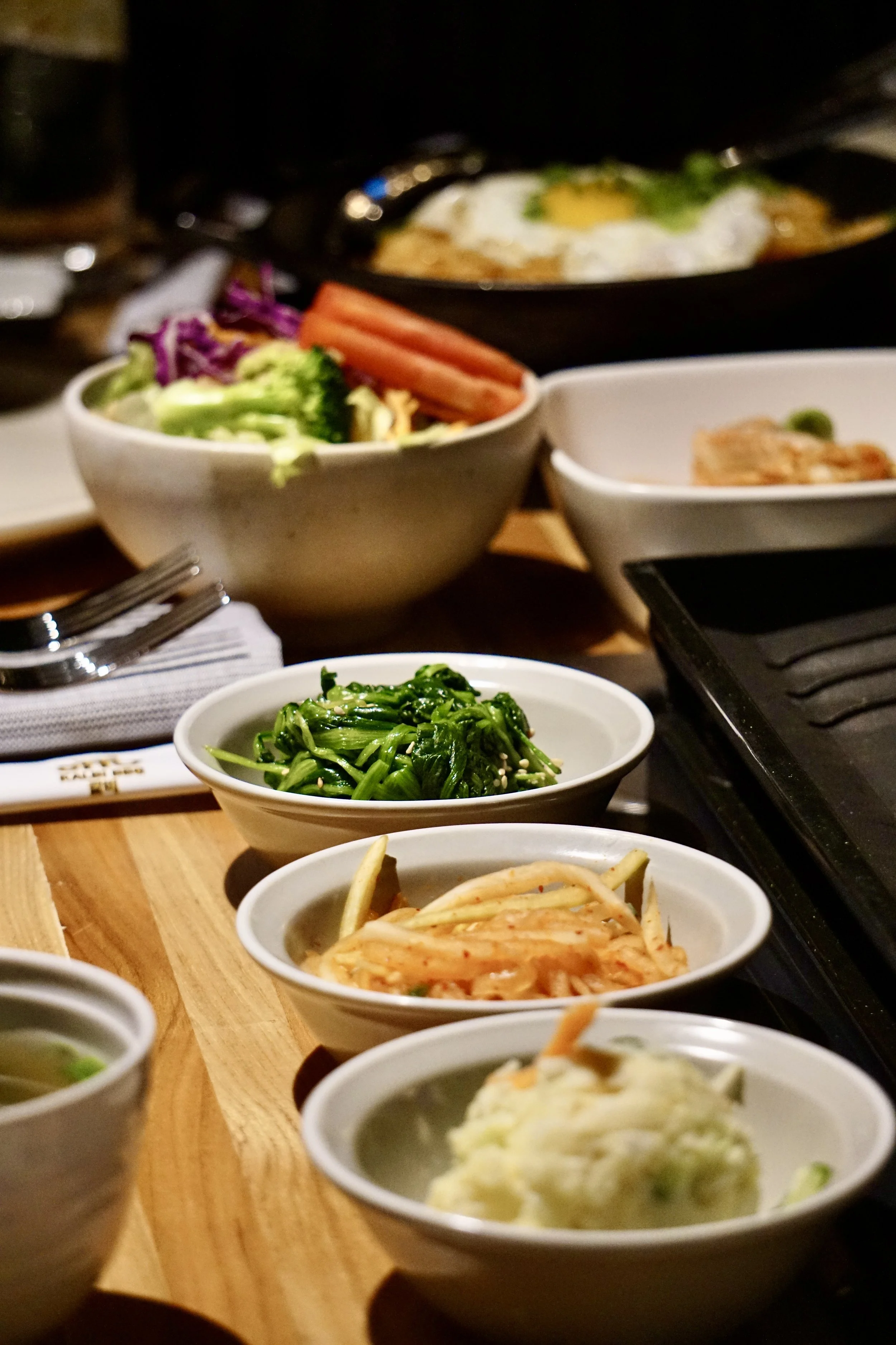 Bowl of assorted Korean side dishes including spinach, kimchi, and pickled vegetables on a wooden table at a Korean restaurant.