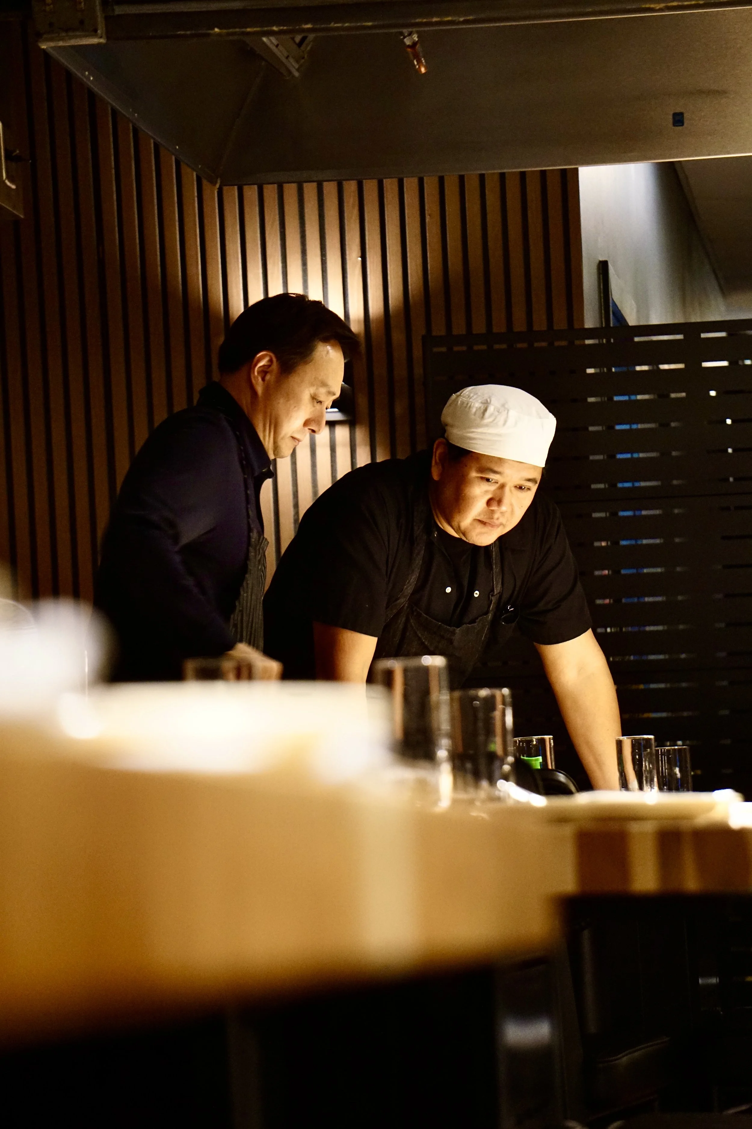 Two chefs working in a restaurant kitchen, one wearing a white chef's hat and the other without a hat, both focusing on their tasks with glasses and dishes on the counter in front of them.