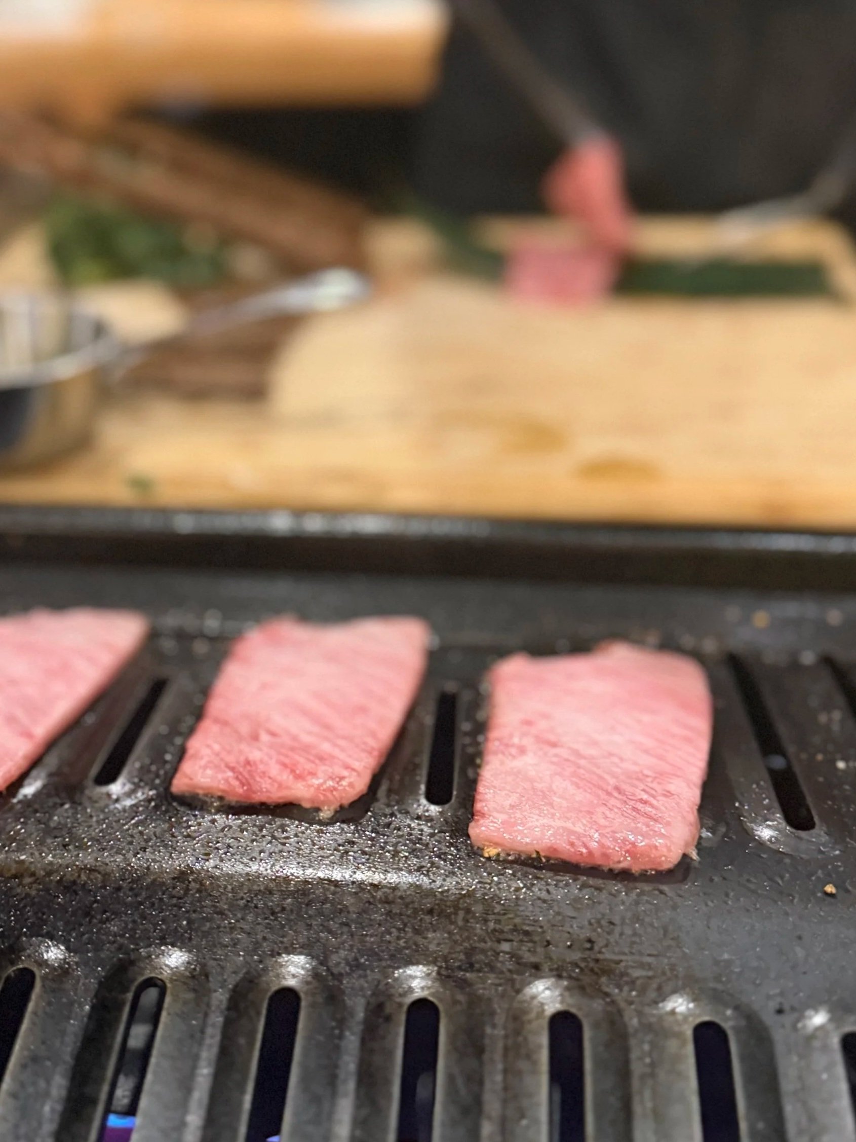 Three pieces of pink meat grilling on a black grill with some juice and seasoning, out of focus cooking surface in the background.