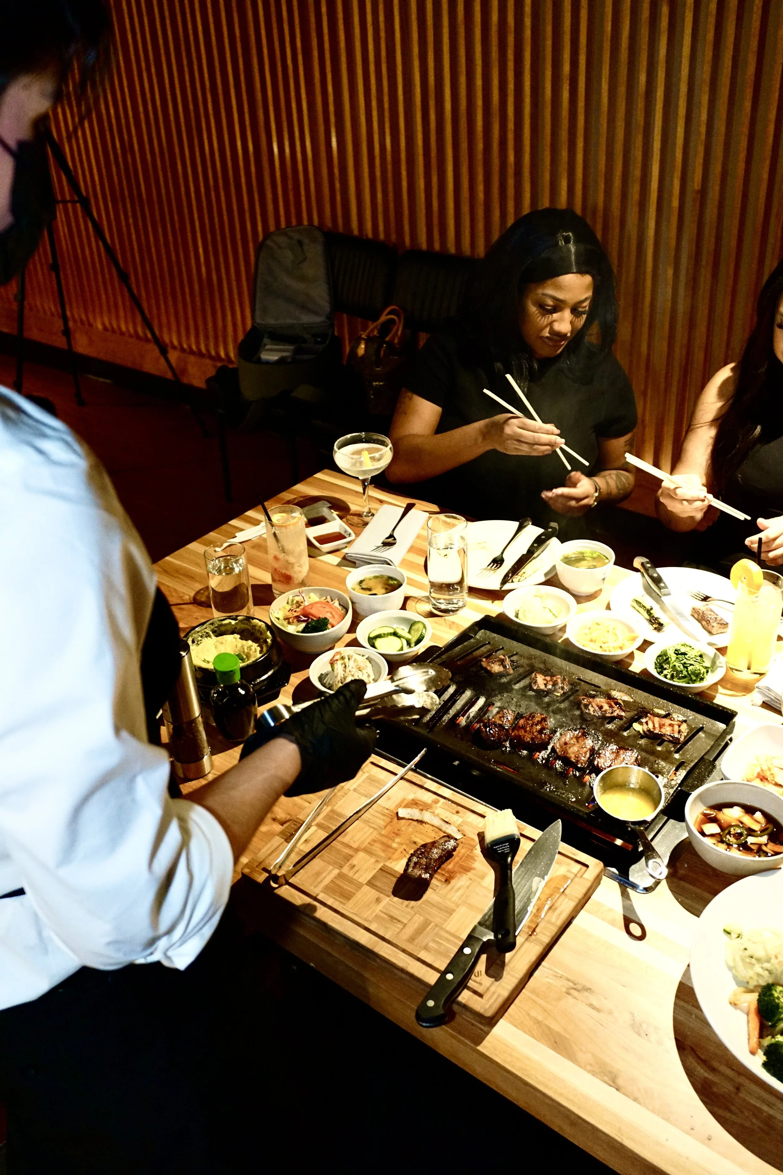 People dining at a restaurant table with various dishes, including grilled meat, bowls of soup, salad, and drinks, while a chef prepares food.