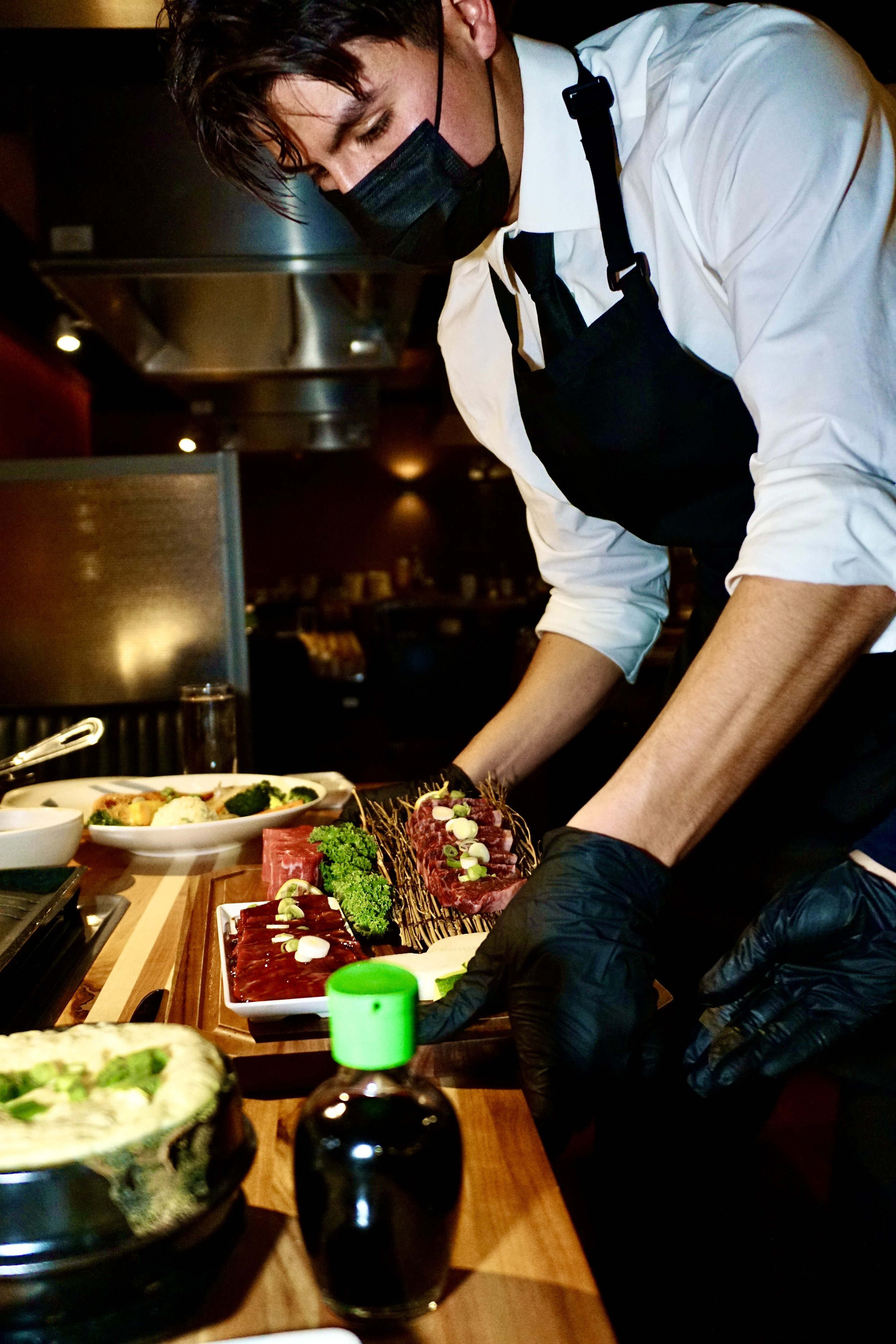 A chef wearing a white shirt, black apron, black face mask, and black gloves is preparing sashimi at a restaurant counter.