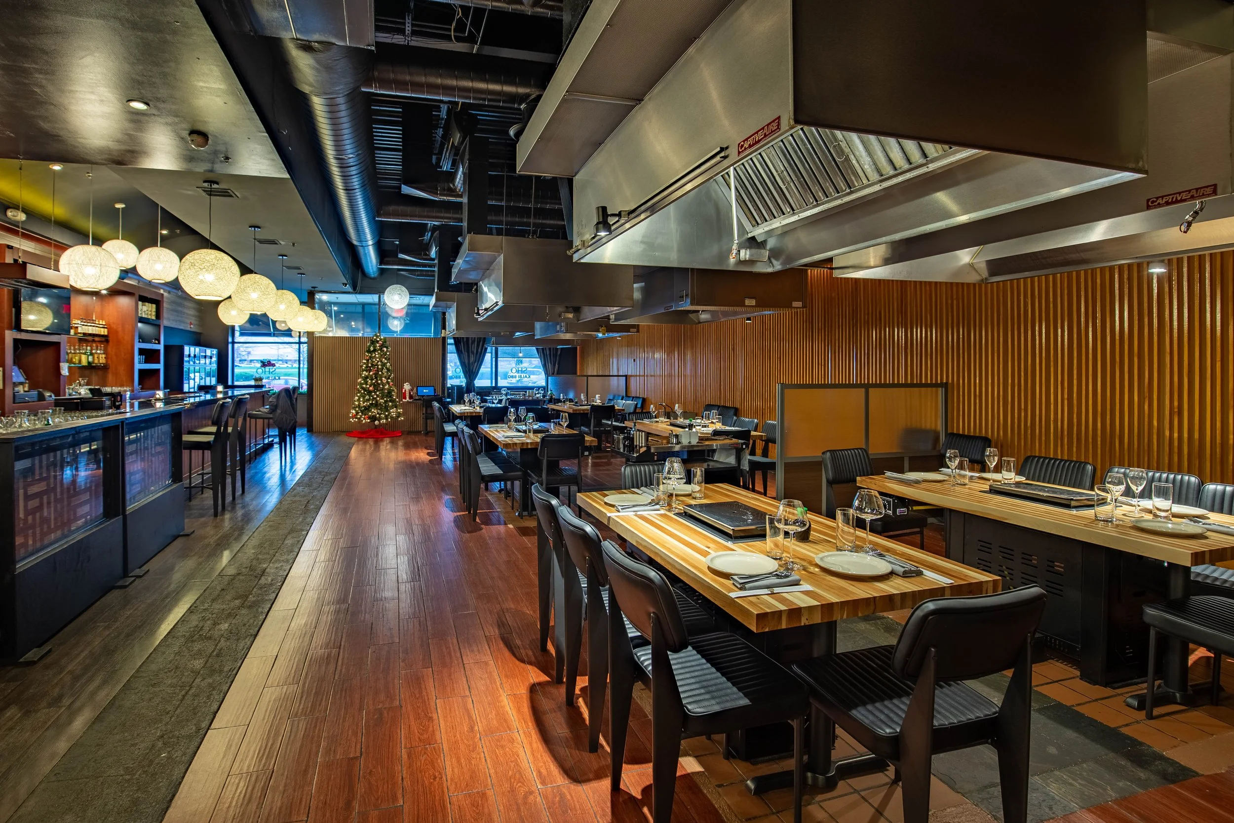 Empty restaurant with wooden tables, black chairs, a decorated Christmas tree near the window, and a bar with hanging spherical lights.