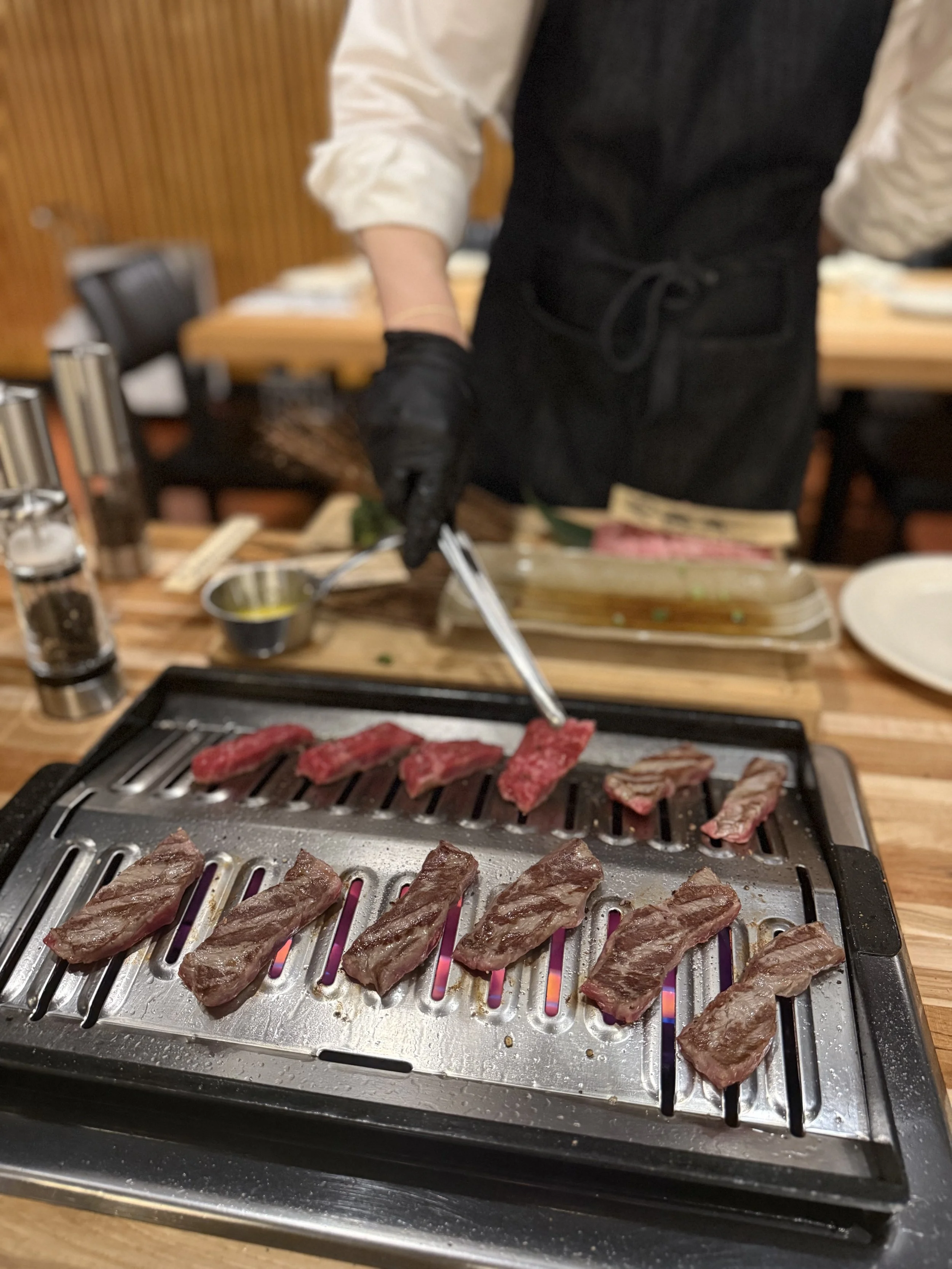 A person grilling slices of beef on a tabletop grill, wearing black gloves and a black apron, with a wooden table and various condiments in the background.