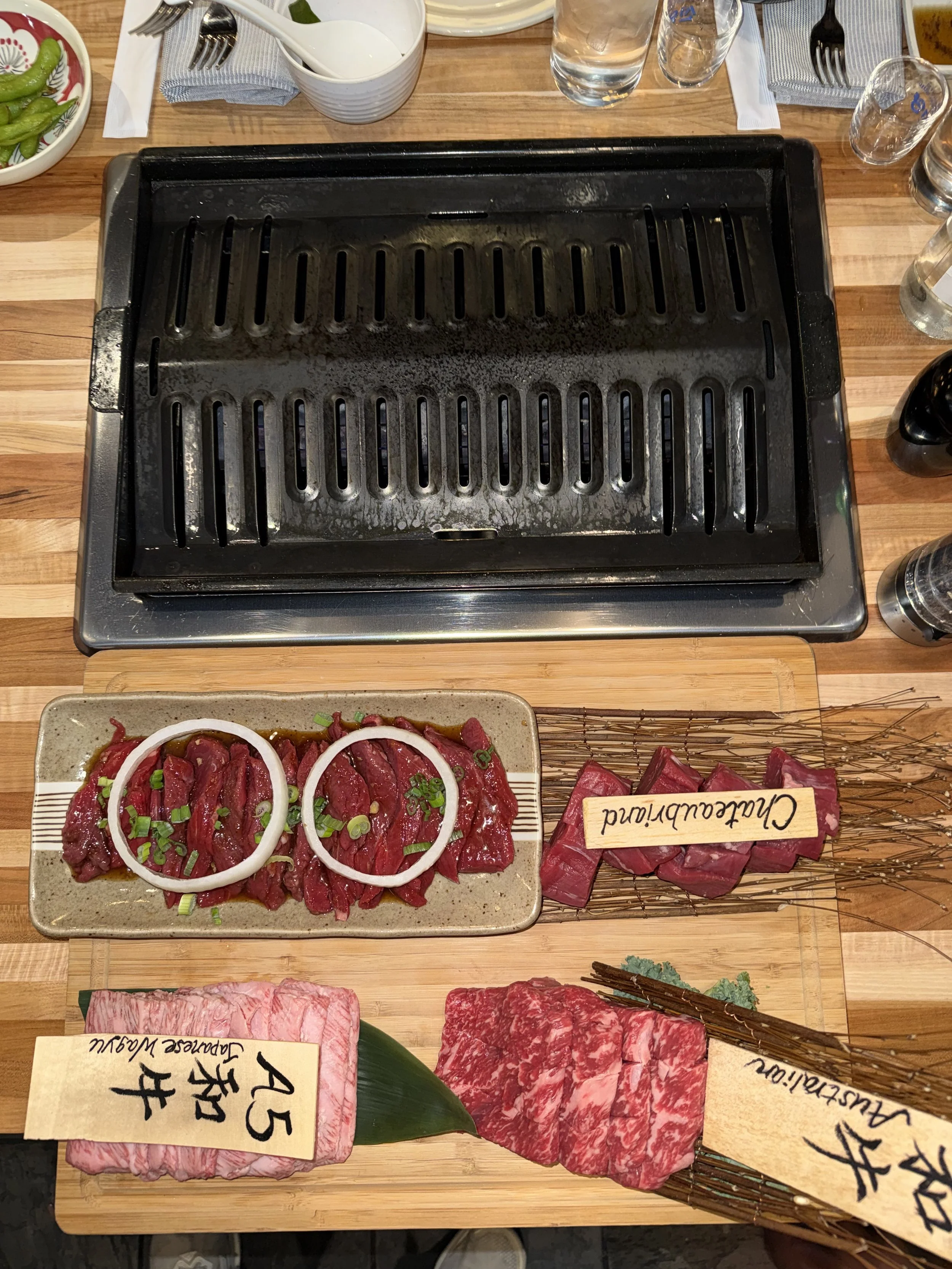 Un grill électrique avec des tranches de viande crues déployées devant. La viande est fraîche et bien marbrée. À côté, il y a une assiette avec de la viande assaisonnée garnie d'oignons et de ciboulette, une étiquette marquée 'Charculand' ou 'Charcul