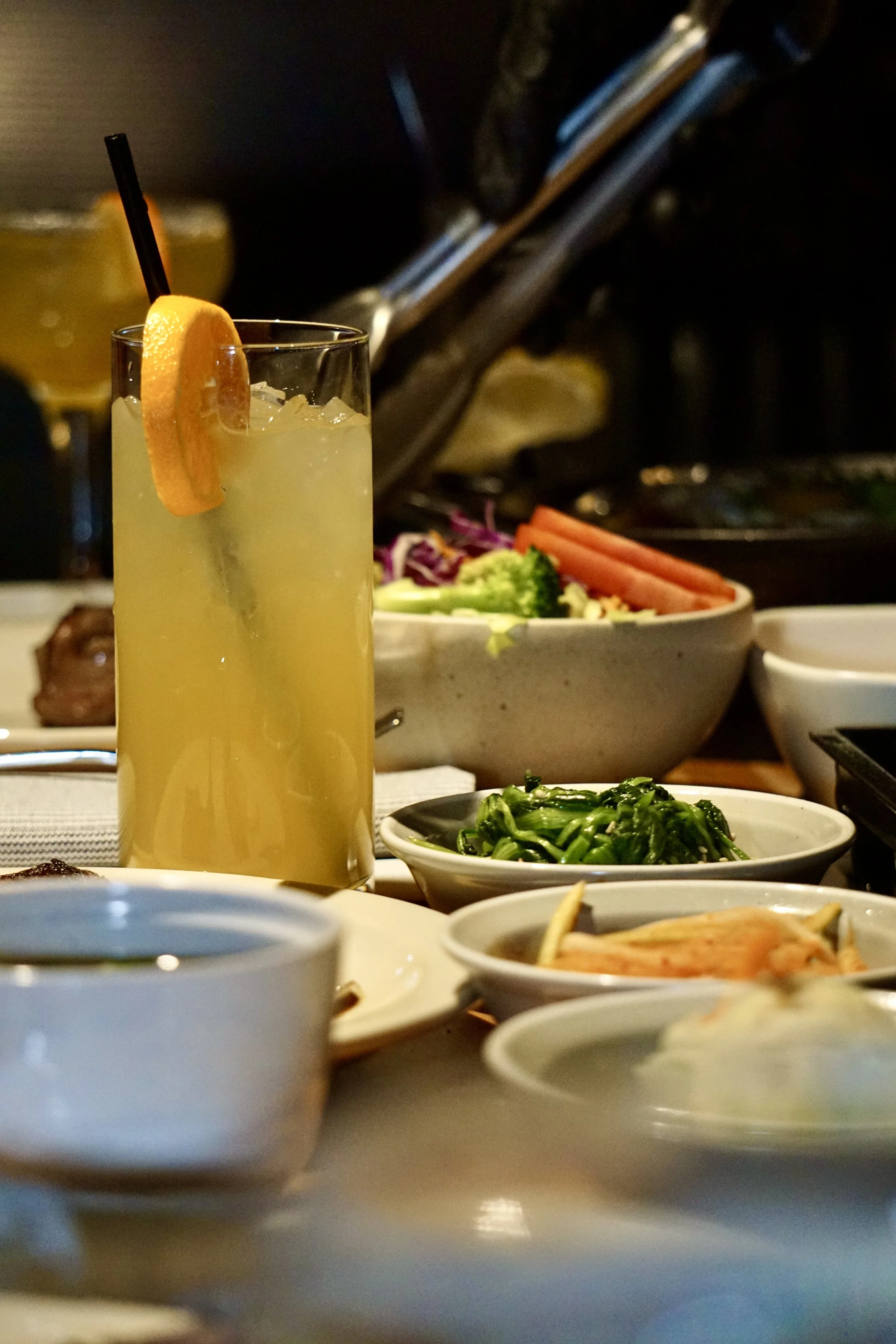 A tall glass of yellow beverage with ice, a lemon wedge, and a straw on a table with various dishes including a salad, vegetables, and small bowls of condiments.