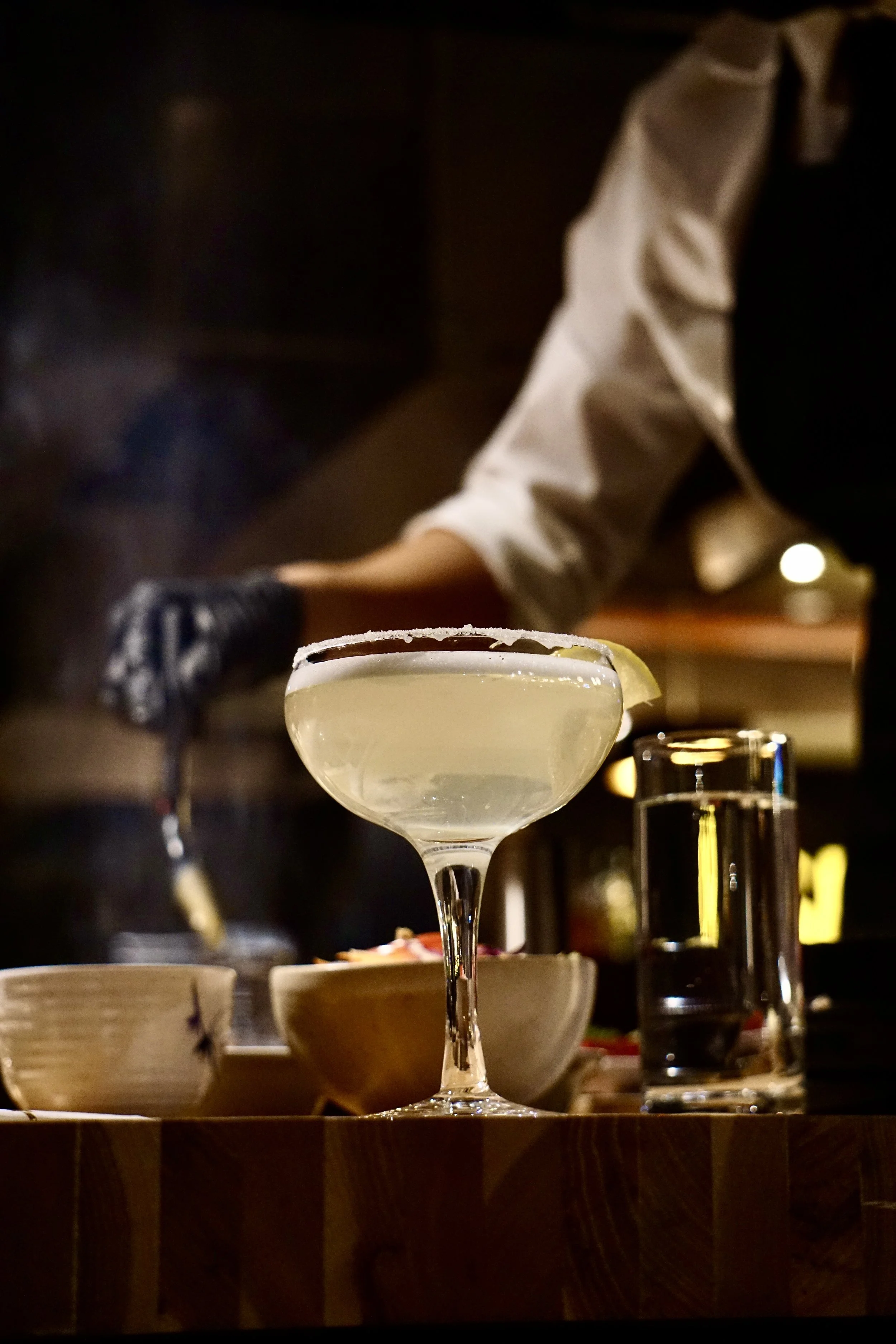 A cocktail glass with a lemon wedge, salt rim, and clear drink, on a wooden table with bowls and a glass of water, with a bartender in the background.