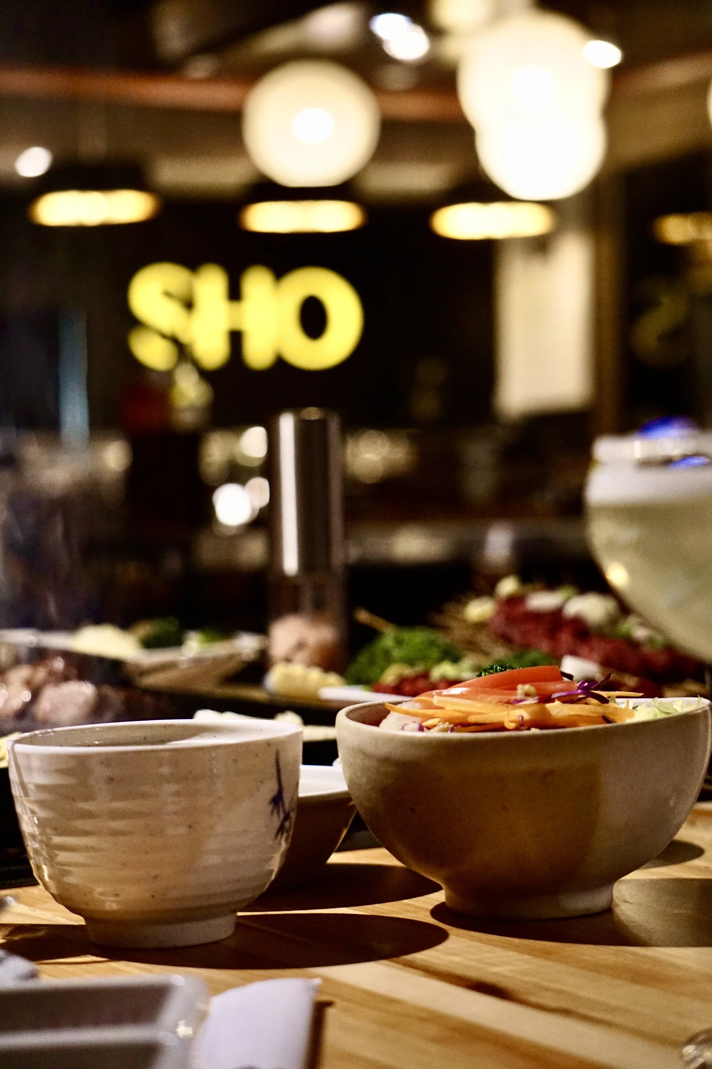 Close-up of a bowl of salad and a small bowl on a wooden table in a restaurant with a blurred background and a yellow illuminated 'SHO' sign.