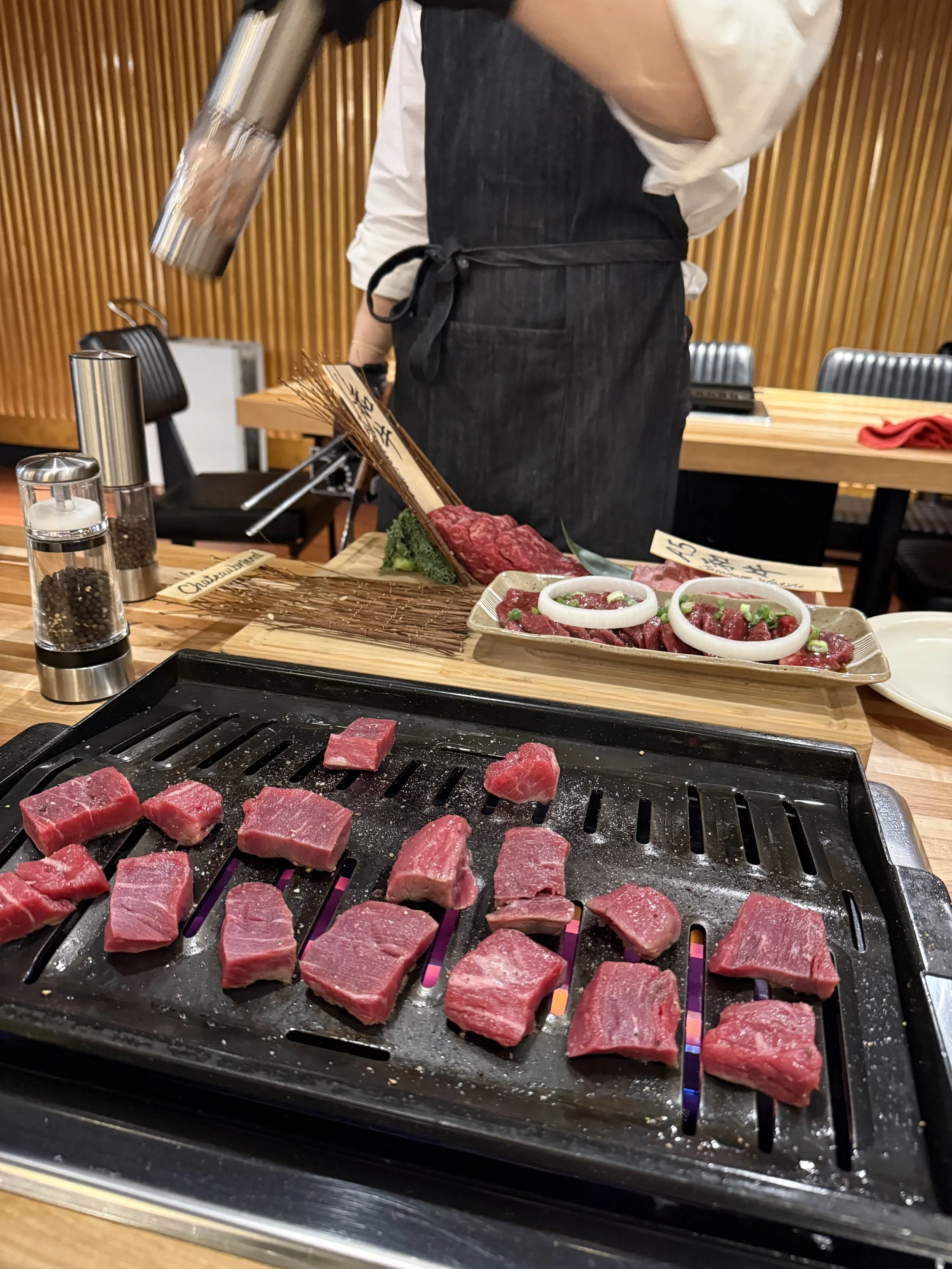 Cooking meat on a grill at a restaurant, with a chef or waiter preparing ingredients in the background.