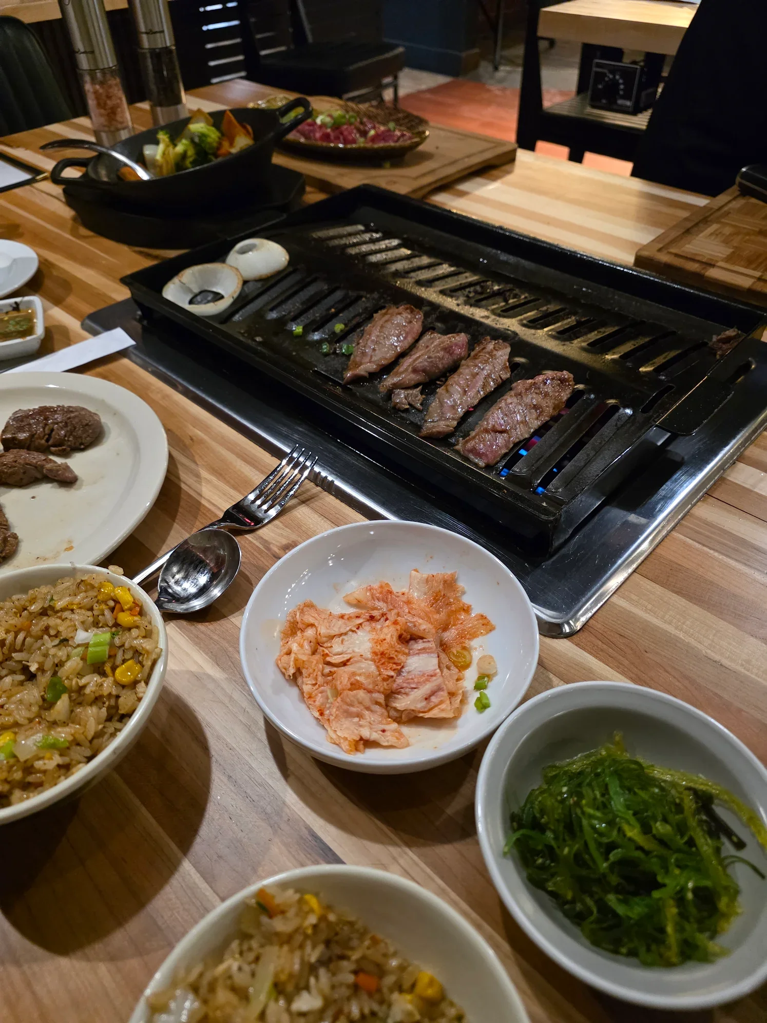 A table set for Korean barbecue with a tabletop grill containing slices of beef, onions, and green onions, surrounded by assorted dishes including kimchi, rice, seaweed salad, and small bowls of sauces.