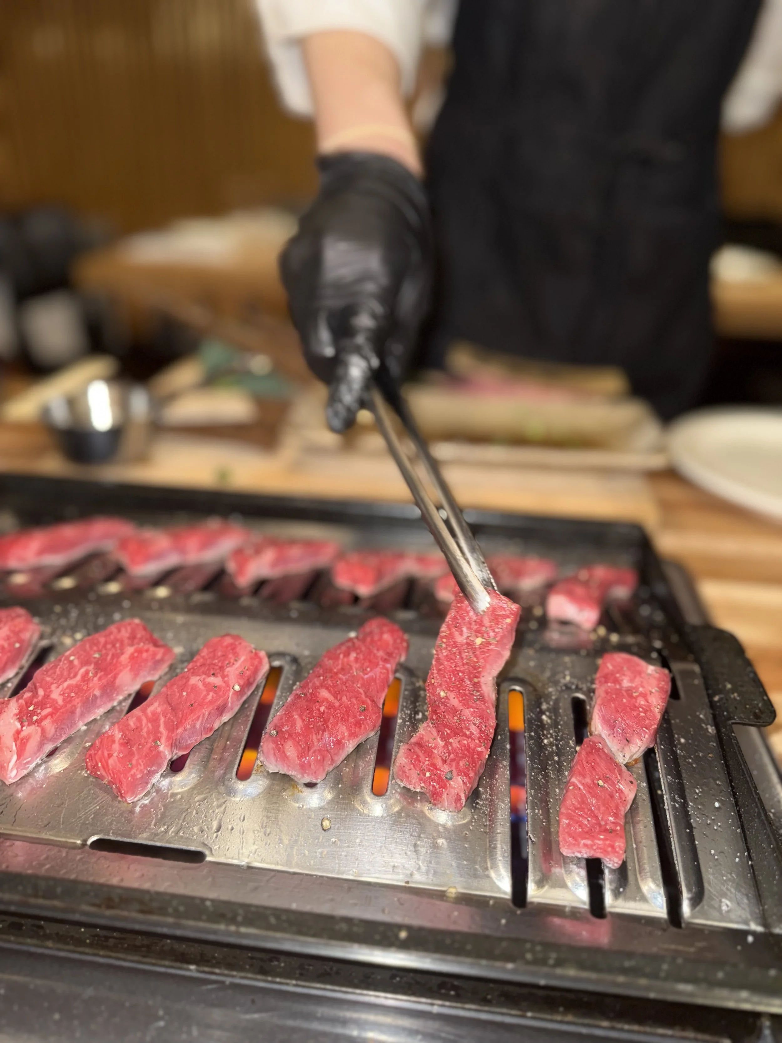 Person using tongs to cook strips of beef on a grill.