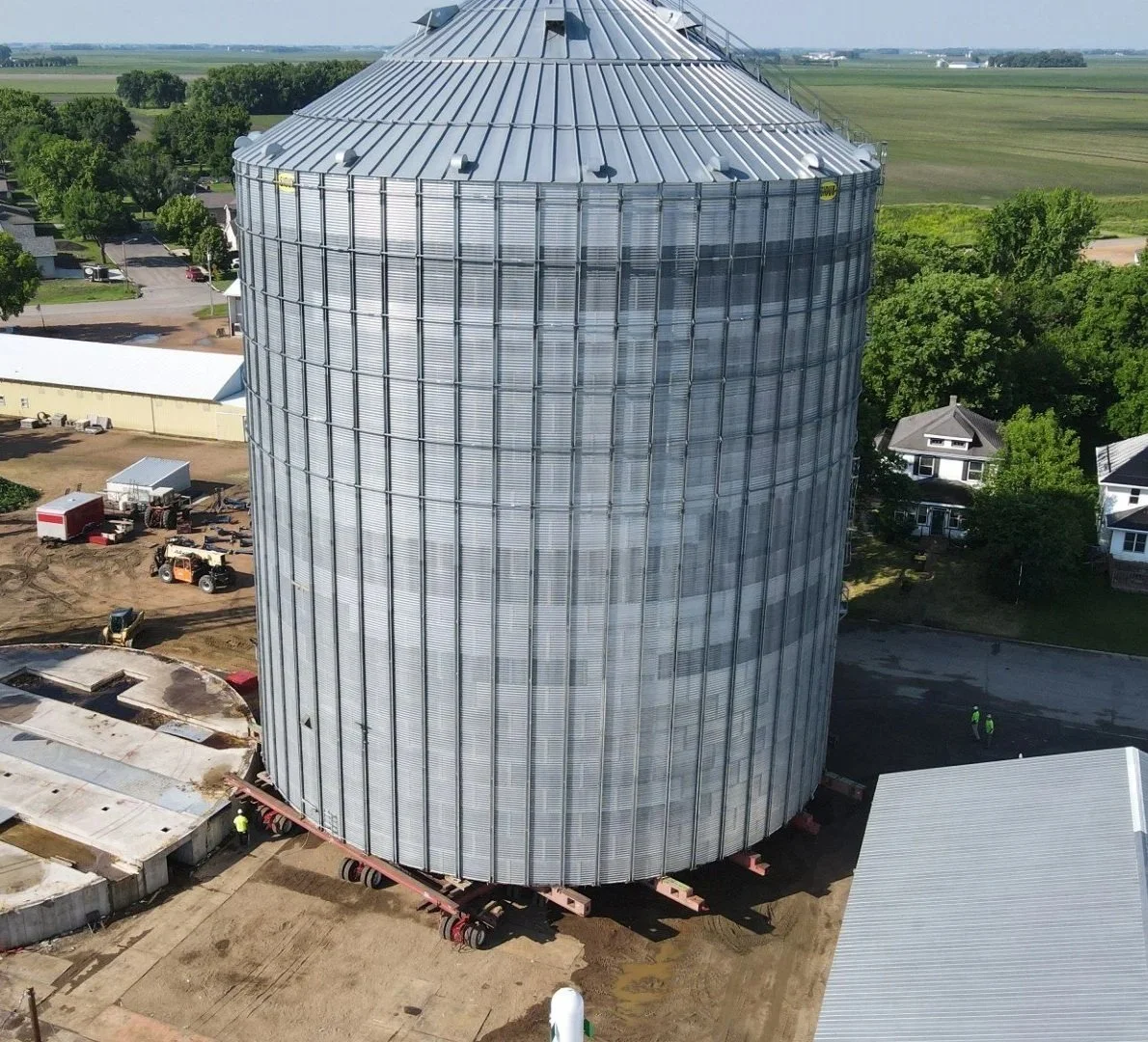 Construction site with a large metal grain bin being relocated, surrounded by construction equipment, workers, and nearby houses and fields.