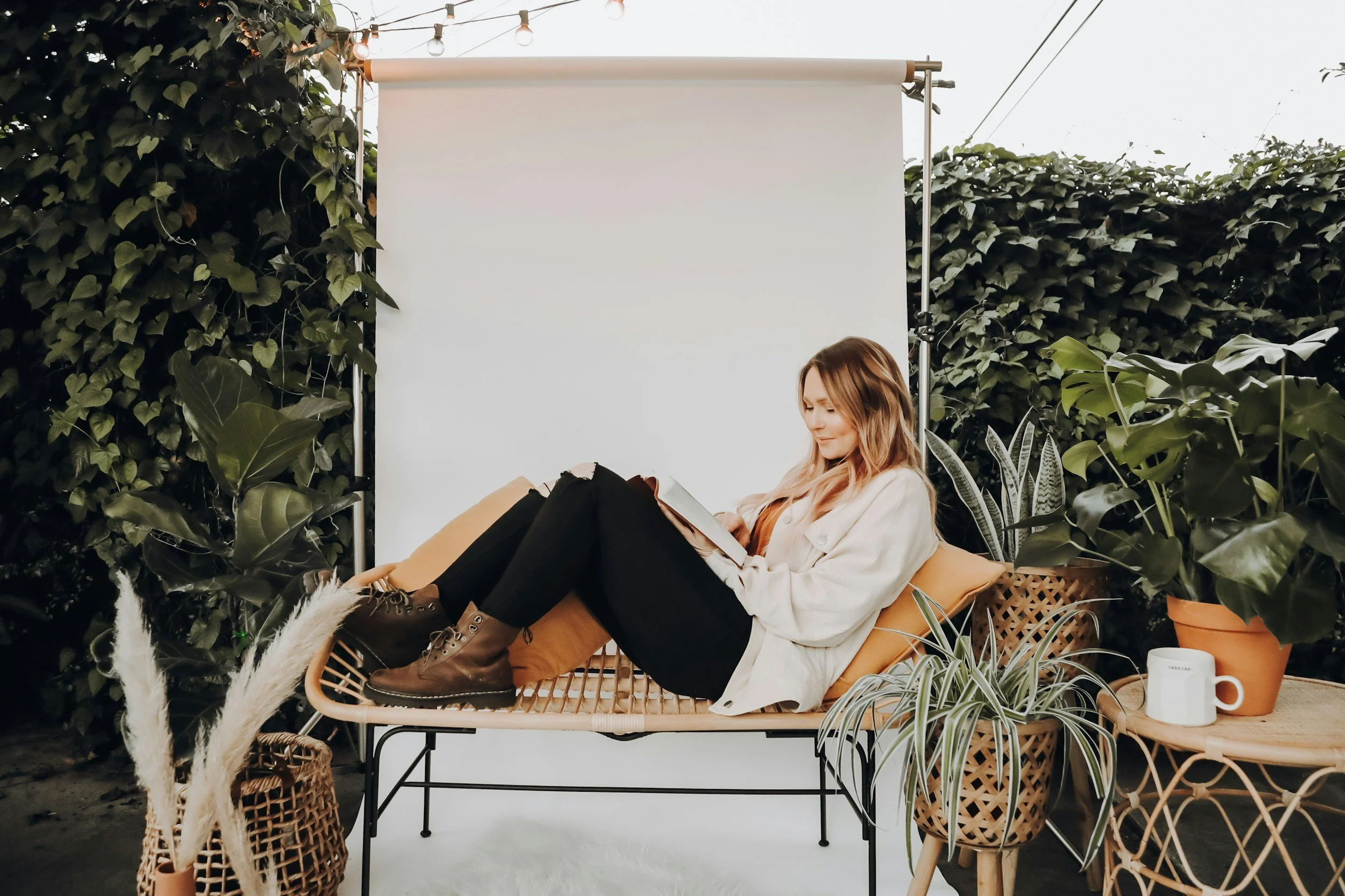 A woman relaxing on a wicker bench outdoors, reading a book, surrounded by potted plants and outdoor decor, with a white backdrop and string lights overhead.