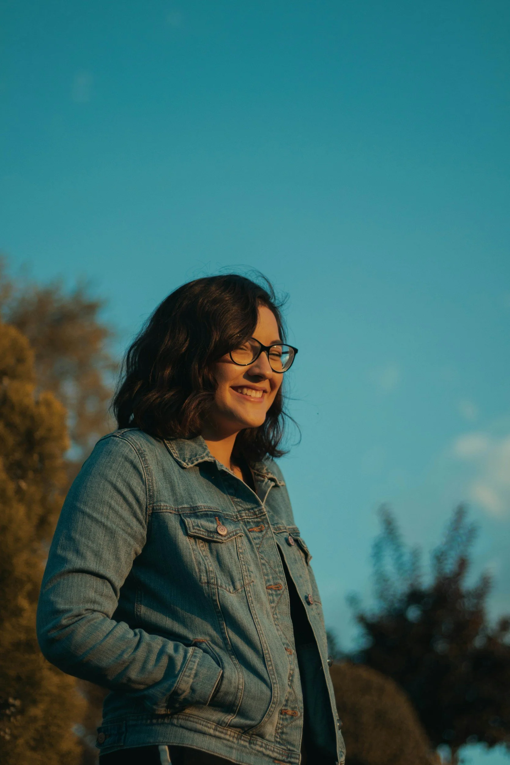 A young woman with glasses and dark wavy hair smiling with eyes closed, wearing a denim jacket outdoors during golden hour, with trees and sky in the background.