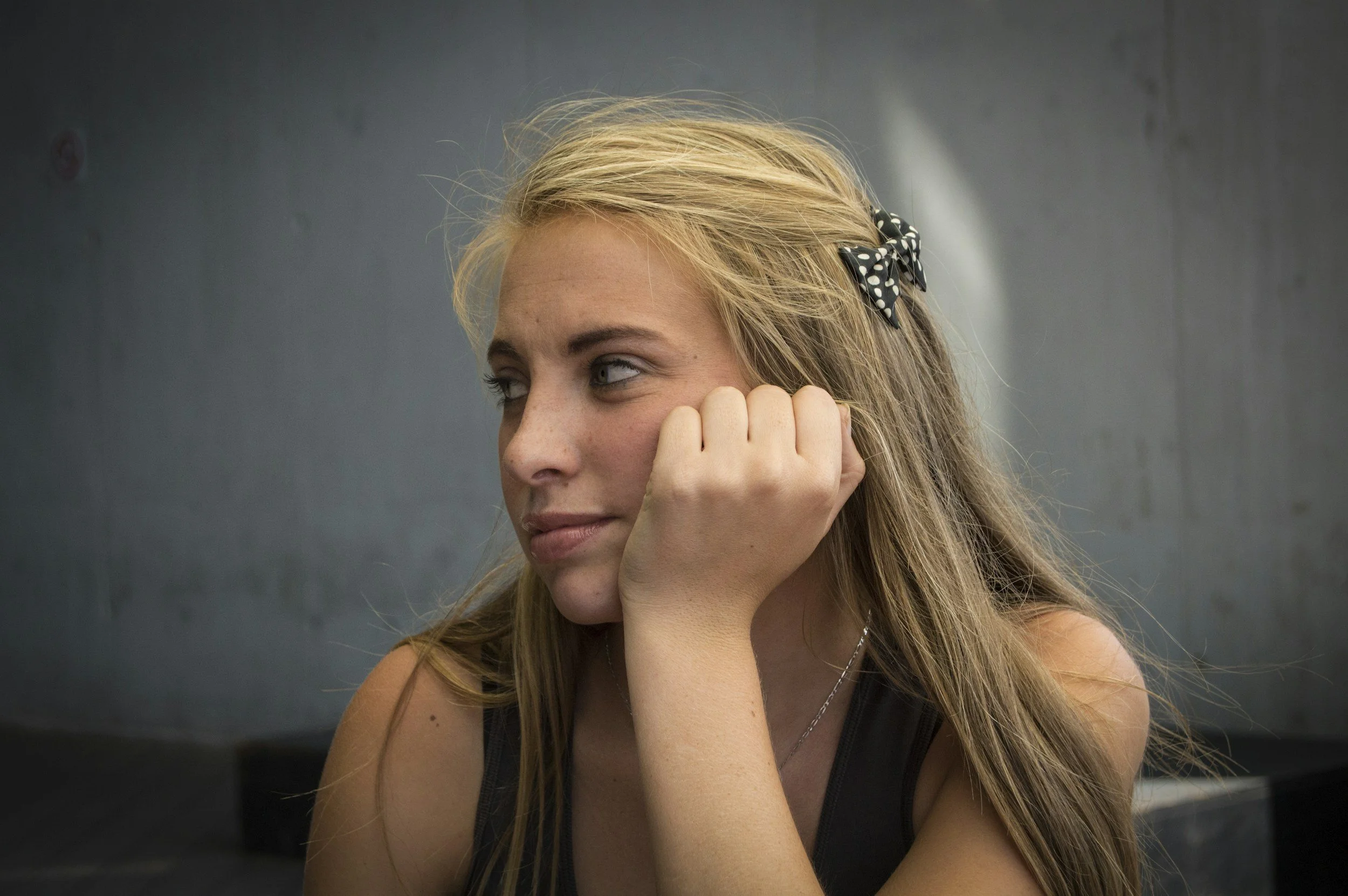 A young woman with blonde hair styled with a black and white polka dot bow, resting her chin on her hand and looking to the side, sitting against a gray wall.