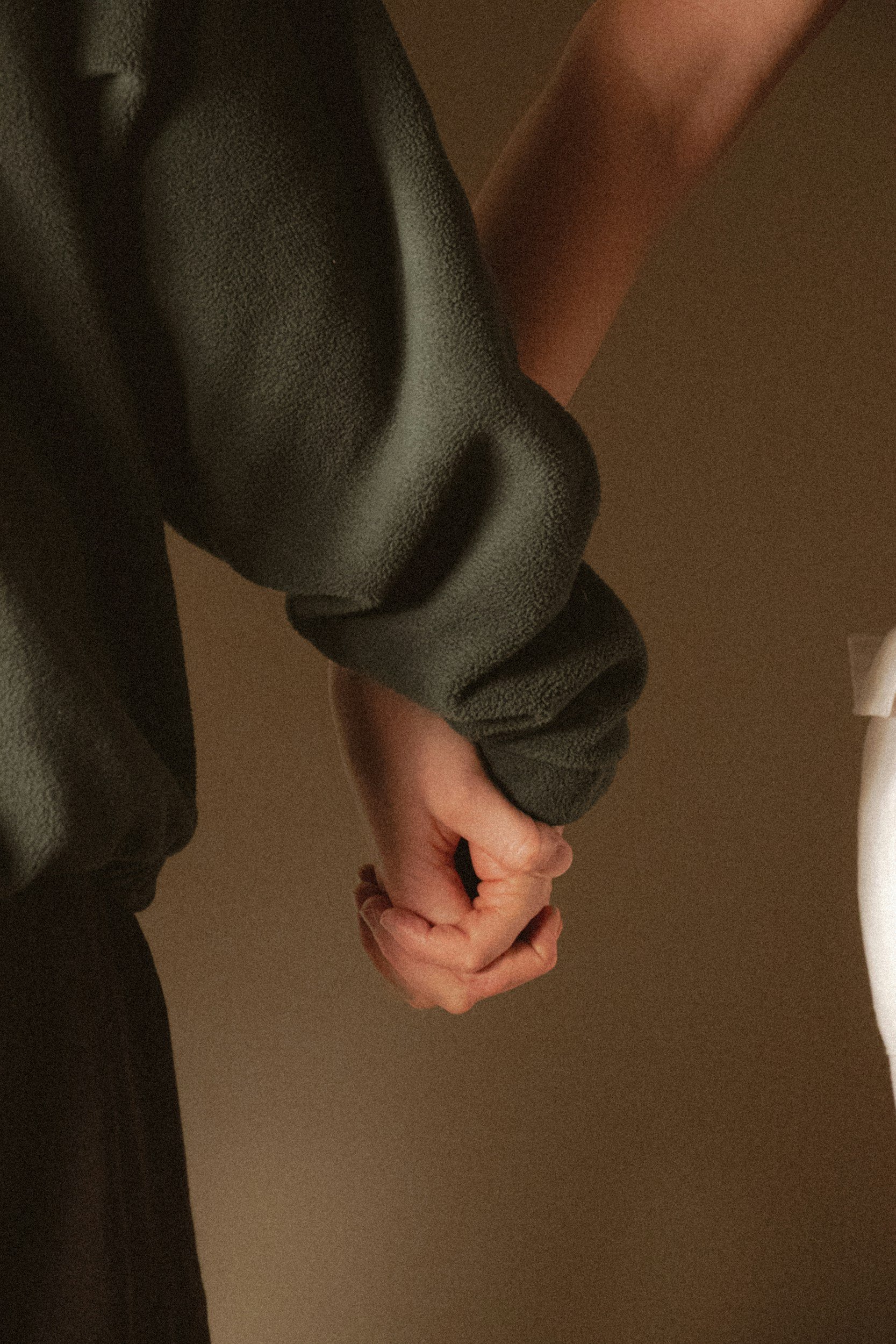 A person wearing a dark hoodie is holding their hands together with fingers intertwined, standing indoors near a brown wall and a white object.