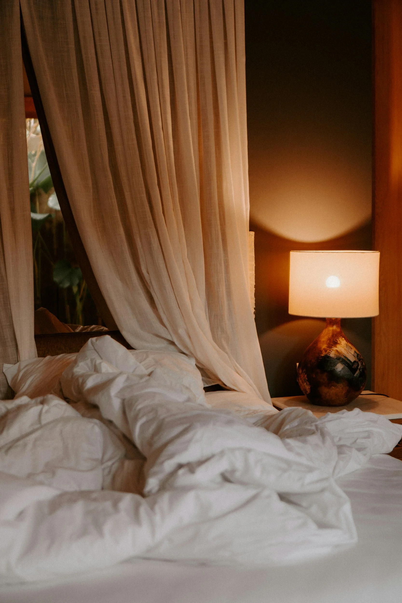 Unmade bed with white sheets and pillows, a cream-colored curtain, and a bedside table with a ceramic lamp in a dimly lit room.