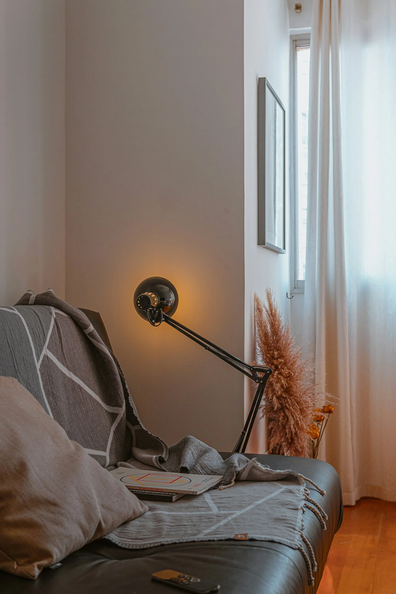 A cozy living room corner with a sofa, throw pillows, a blanket, a modern black adjustable desk lamp, a magazine, and a remote on the sofa. There is a window with white curtains letting in natural light, and decorative dried plants near the window.