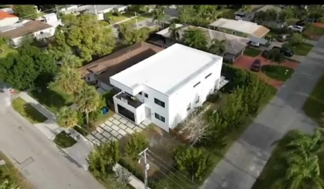 Aerial view of a modern white two-story house with a flat roof, surrounded by a well-maintained yard and neighboring houses in a suburban neighborhood.