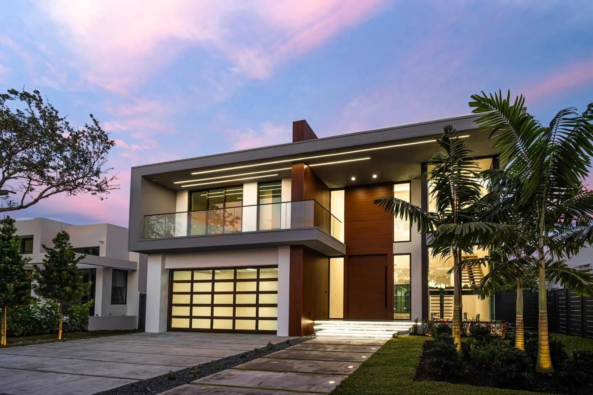 Modern two-story house with large glass windows, a double garage with frosted glass doors, a small front yard with palm trees, and steps leading to the entrance, illuminated at dusk with a colorful sunset sky in the background.